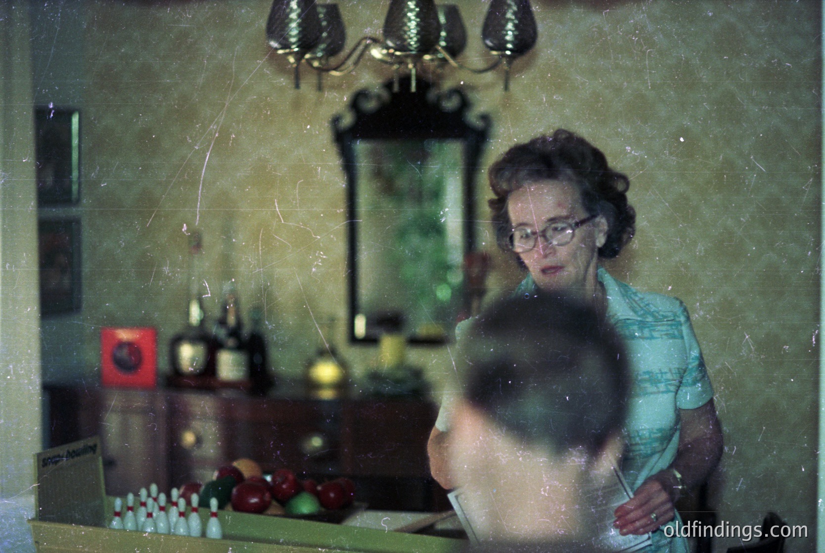 Vintage indoor scene featuring a woman in a light blue blouse standing beside a kitchen countertop adorned with glass bottles, a bowl of fruit, and a red sign. Reflections in a mirror reveal a chandelier and a blurred figure. Mid-20th century domestic interior, likely European.