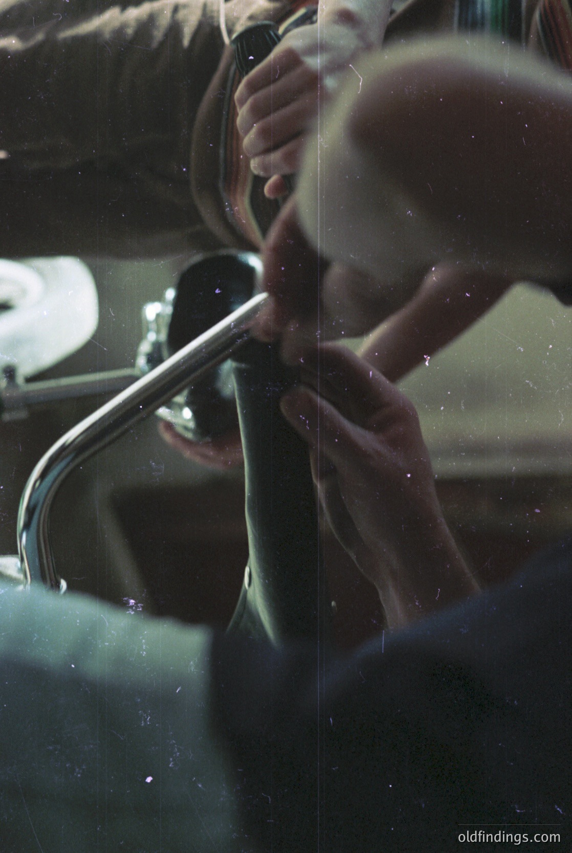 Close-up of a person adjusting a vintage car’s chrome fender mirror, visible through a rain-spotted windshield. Reflections and condensation blur background details, emphasizing the mirror’s polished surface and the driver’s focused hand. Likely mid-20th century automotive design.