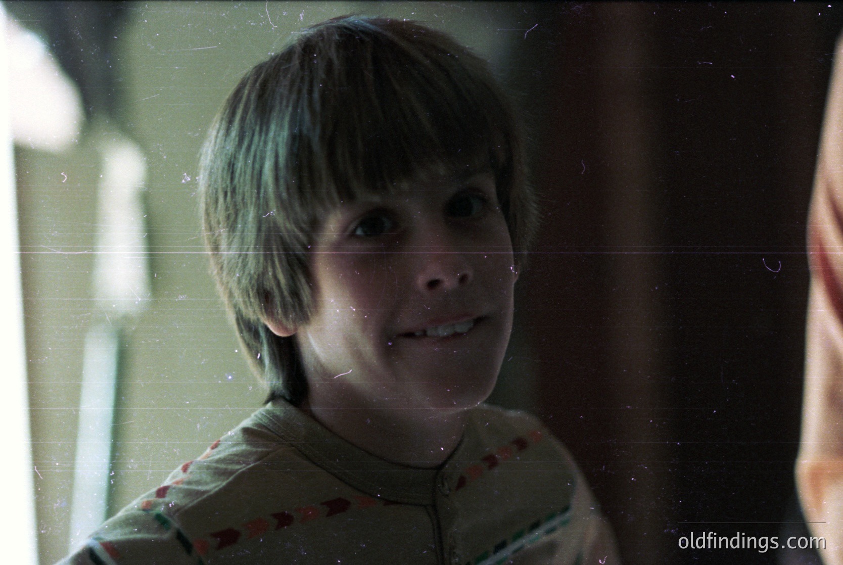 Vintage-style close-up of a young boy with short, parted hair, smiling slightly. He wears a floral-patterned shirt with red accents. Blurred background suggests indoor lighting, likely from a window or lamp. Style evokes mid-20th century family photography ().
