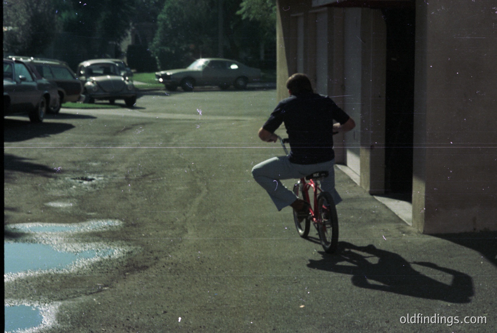 Mid-20th century street scene with a cyclist in motion. The person wears a dark jacket, light pants, and a cap, riding a red-framed bike. Parked vintage cars (1960s-70s) line the road, and a concrete sidewalk with puddles reflects sunlight. Urban residential area with trees and a building in the background.