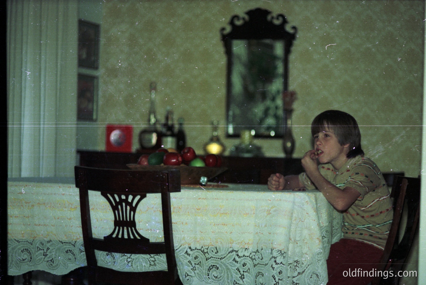 Vintage indoor scene featuring a child seated at a wooden table draped with a lace tablecloth. Reflective mirror reveals a framed mirror, bottles, and a red object on a shelf. Mid-century domestic decor with floral-patterned shirt. Likely Eastern European, 1960s-1970s.