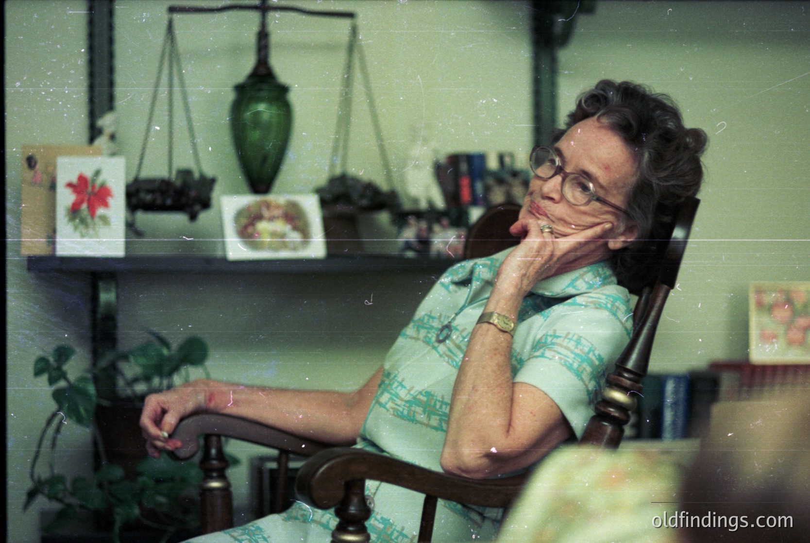 Mid-century woman in floral blouse poses thoughtfully in vintage armchair, surrounded by retro decor: scales, framed botanical art, and potted plants. Lighting suggests indoor setting, likely residential, with muted pastel walls. Style evokes 1960s–1970s domestic life.