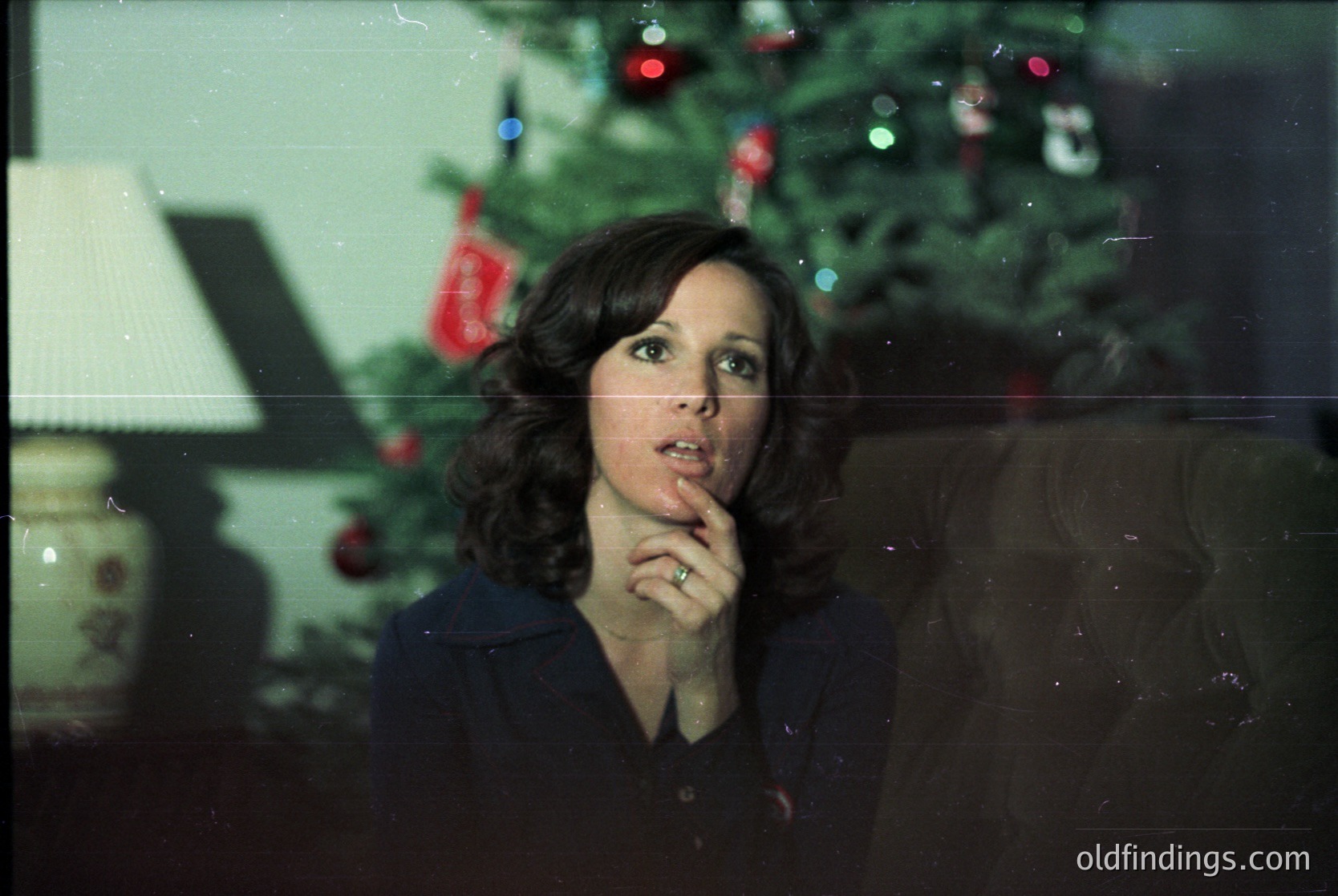 Mid-century woman in a dark blouse poses thoughtfully indoors beside a decorated Christmas tree with red baubles and tinsel, 1970s style. Vintage lighting and floral-patterned lamp add nostalgic warmth.
