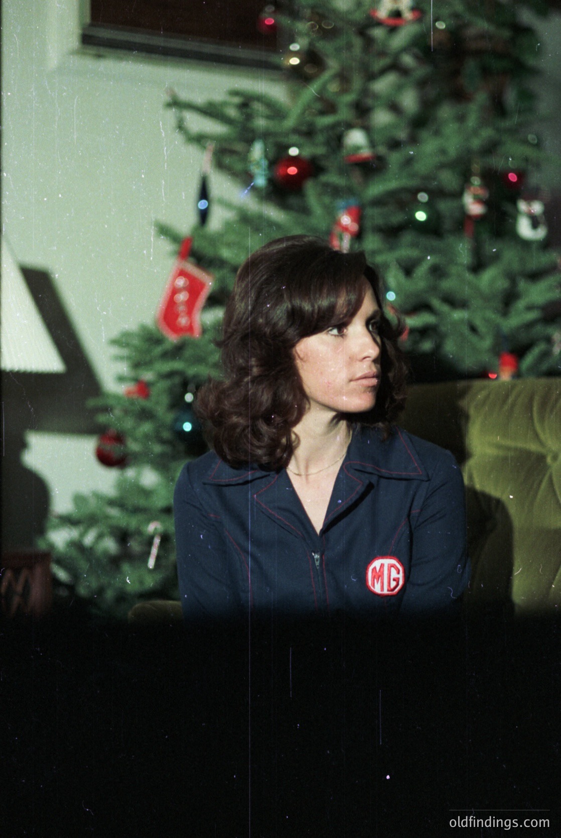A woman in a dark blue jacket with a red "MG" logo poses beside a decorated Christmas tree, likely from the 1960s–1970s. The tree features red baubles, tinsel, and a star topper, suggesting a festive indoor setting.