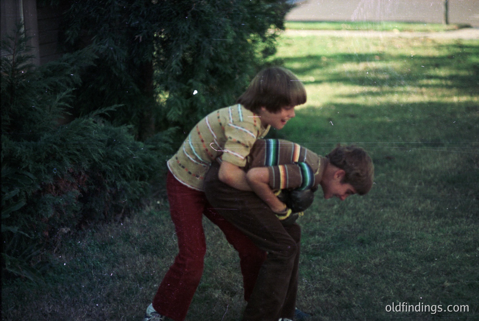 Two boys in 1970s-style clothing play outdoors in a residential yard. The boy on left wears a striped sweater and maroon pants; the other, a striped shirt and brown pants. Lush greenery and a house in background suggest suburban setting.