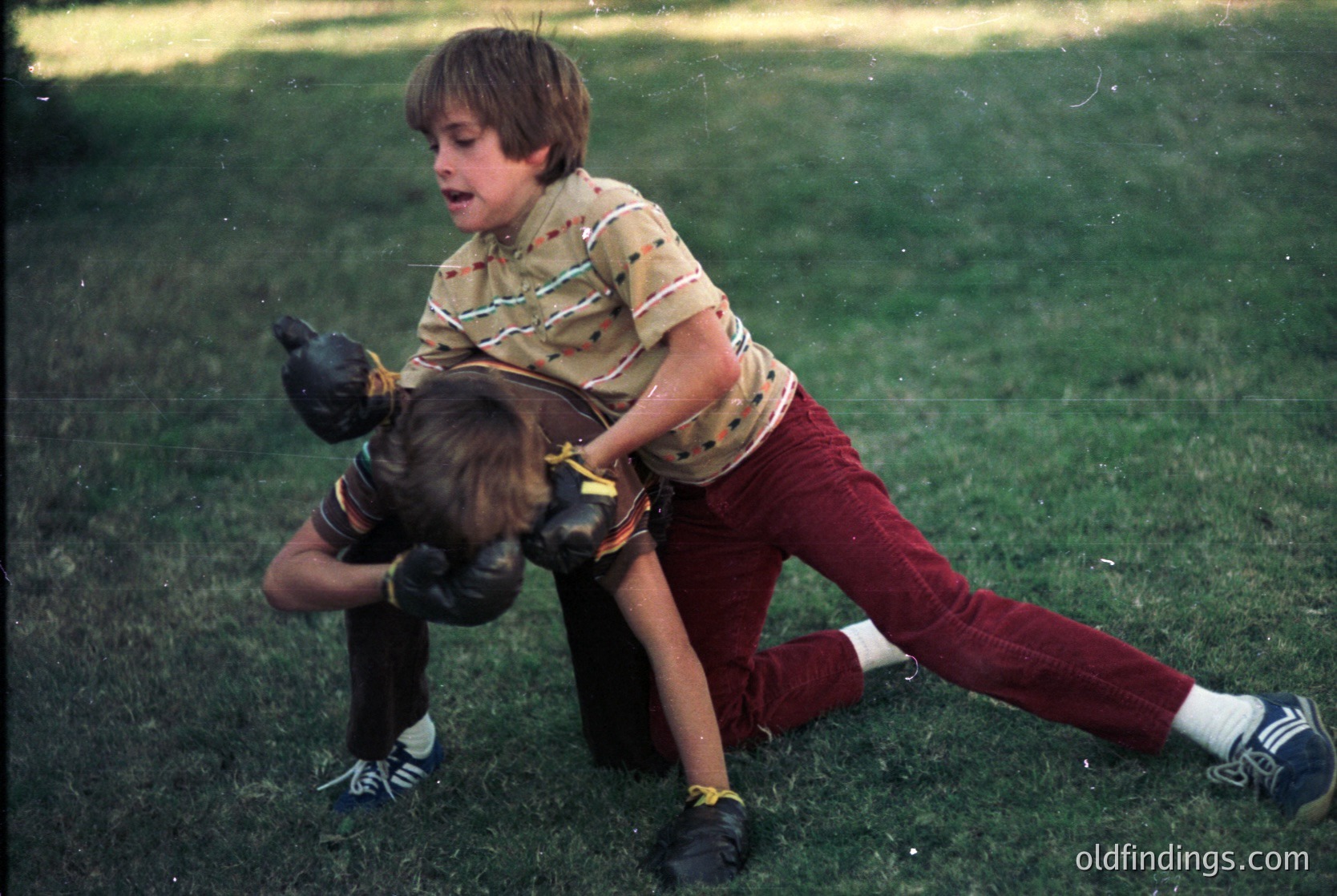Two boys wrestling on grassy field, 1970s-era clothing: striped polo shirt, maroon pants, white socks with stripes. Candid childhood play captures raw energy.