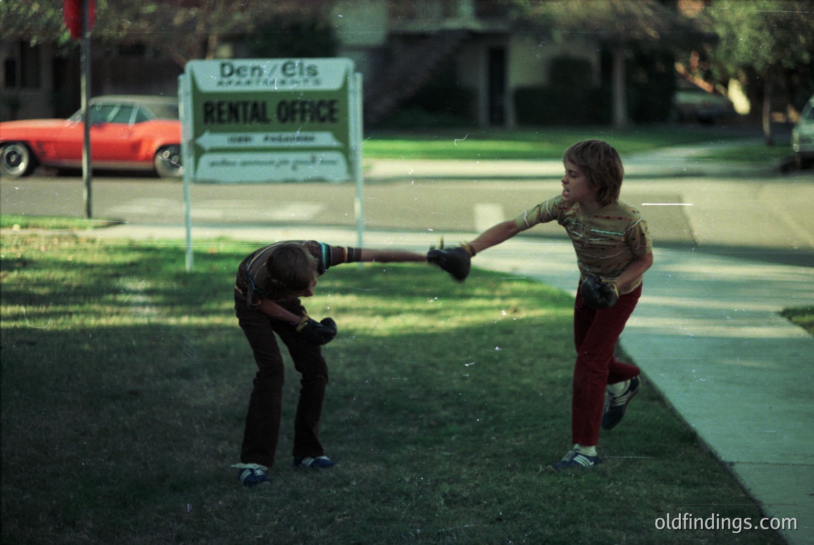Two children playing baseball in a residential yard, 1970s USA. Boy in red pants extends bat while kneeling child swings. "Den Els Rental Office" sign in background. Classic vintage sports scene.