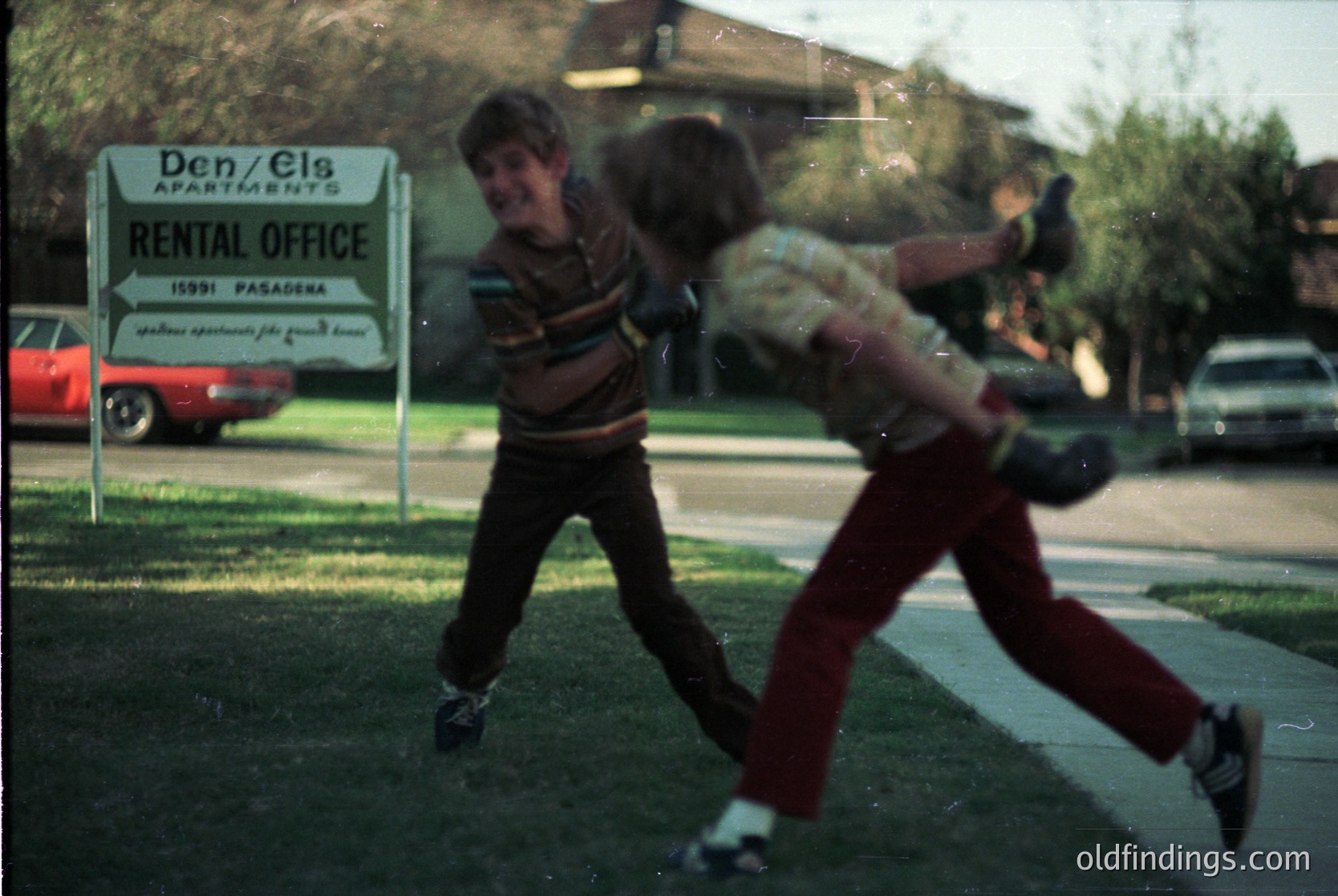 Vintage 1970s Pasadena street scene: two boys mid-play with a baseball in front of a "Den/Els Apartments Rental Office" sign. Classic 70s cars (red sedan, white station wagon) parked on quiet residential street. Grass, sidewalk, and mid-century architecture visible.