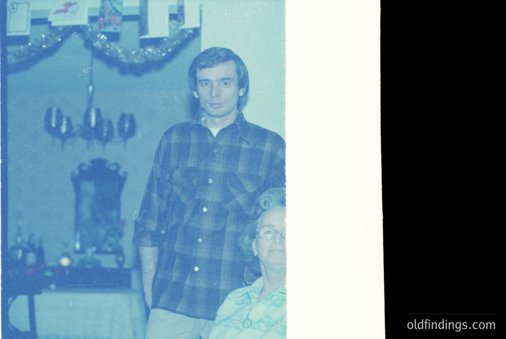 Vintage sepia-toned indoor portrait from the 1970s–1980s, featuring a man in a plaid shirt standing beside an elderly woman in a wheelchair. Decor includes a chandelier, garland, and framed photos. Warm, nostalgic family moment.