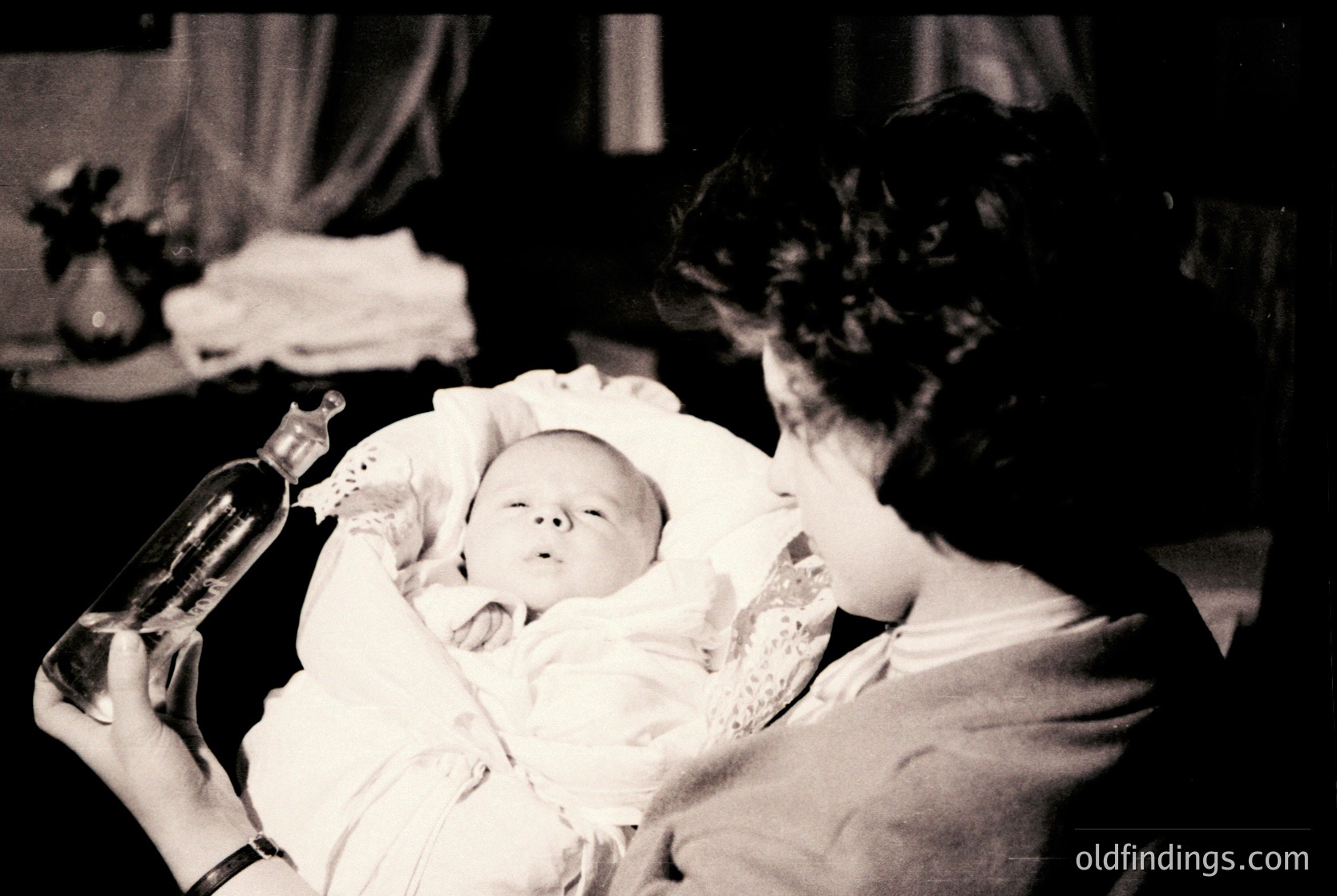 Black-and-white photograph capturing a tender moment: an adult cradles a swaddled infant in a vintage dress, holding a wine bottle in one hand. Soft lighting and blurred background suggest a mid-20th-century domestic setting, likely a 1950s-1960s home.