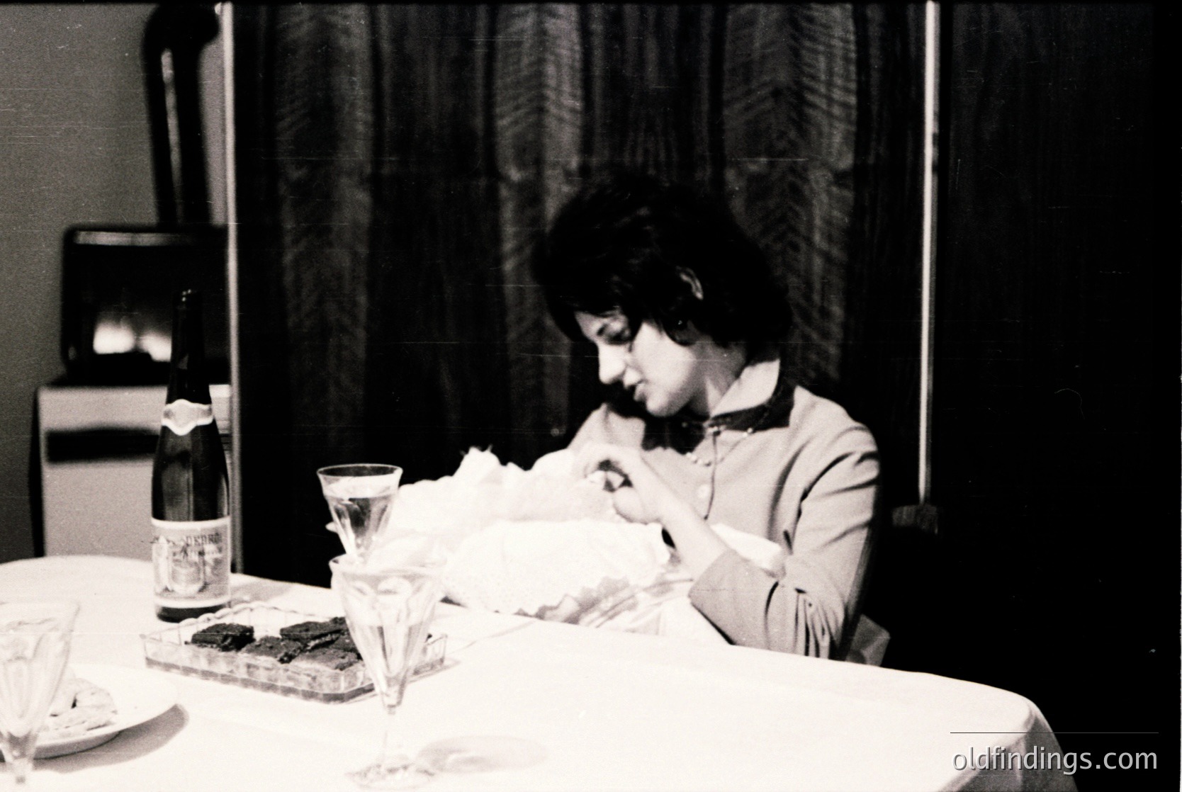 Black-and-white candid of a woman seated at a formal table, engrossed in writing. Mid-20th century attire and vintage typewriter suggest 1950s–1960s domestic or office setting. Empty wine glass, bottle, and plate of food hint at a meal paused for work.