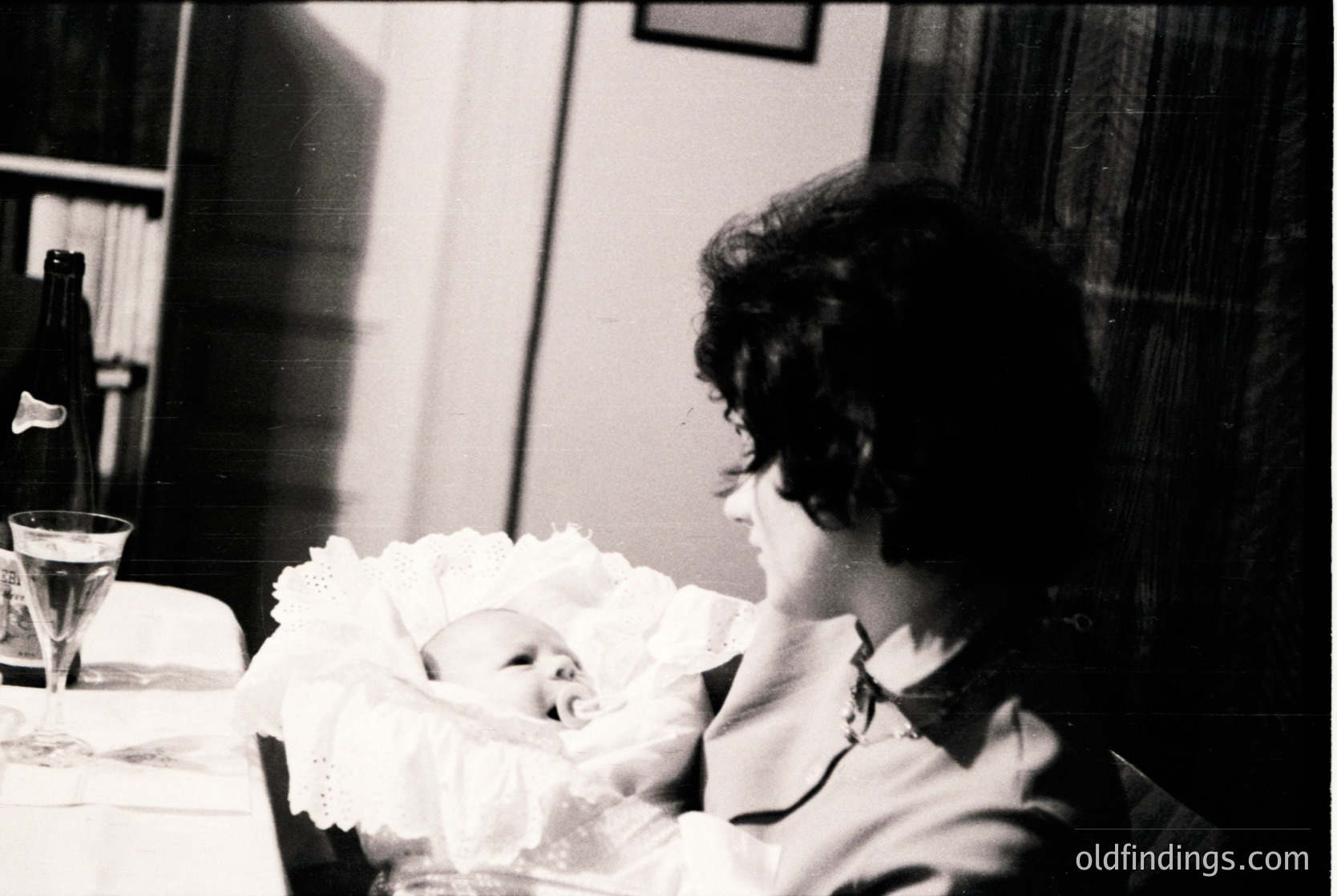 A tender black-and-white moment: a woman cradles a newborn wrapped in a lace-trimmed swaddle, seated at a table with a champagne bottle and flute. Mid-20th century domestic intimacy.
