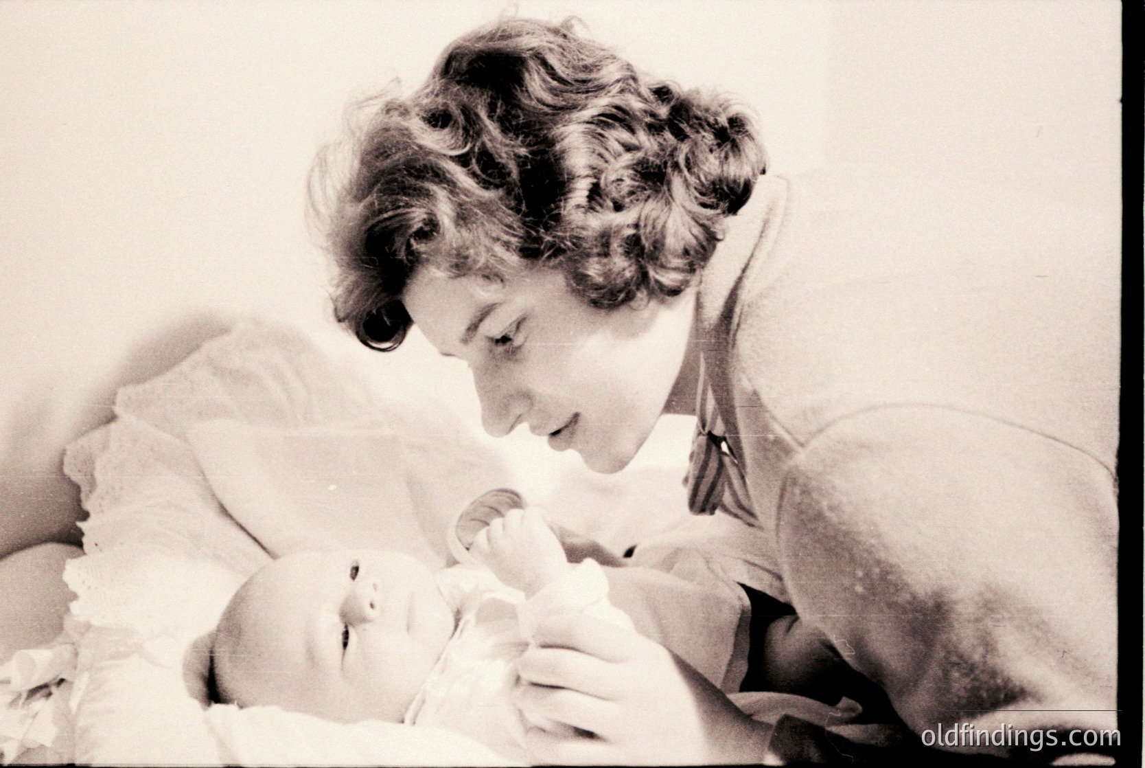 A tender black-and-white portrait of a woman cradling a newborn, likely from the 1950s–1960s. The woman’s hairstyle and clothing—curly hair, a simple blouse, and a brooch—suggest mid-century domestic life. The infant’s swaddling and the woman’s protective posture evoke themes of maternal care.