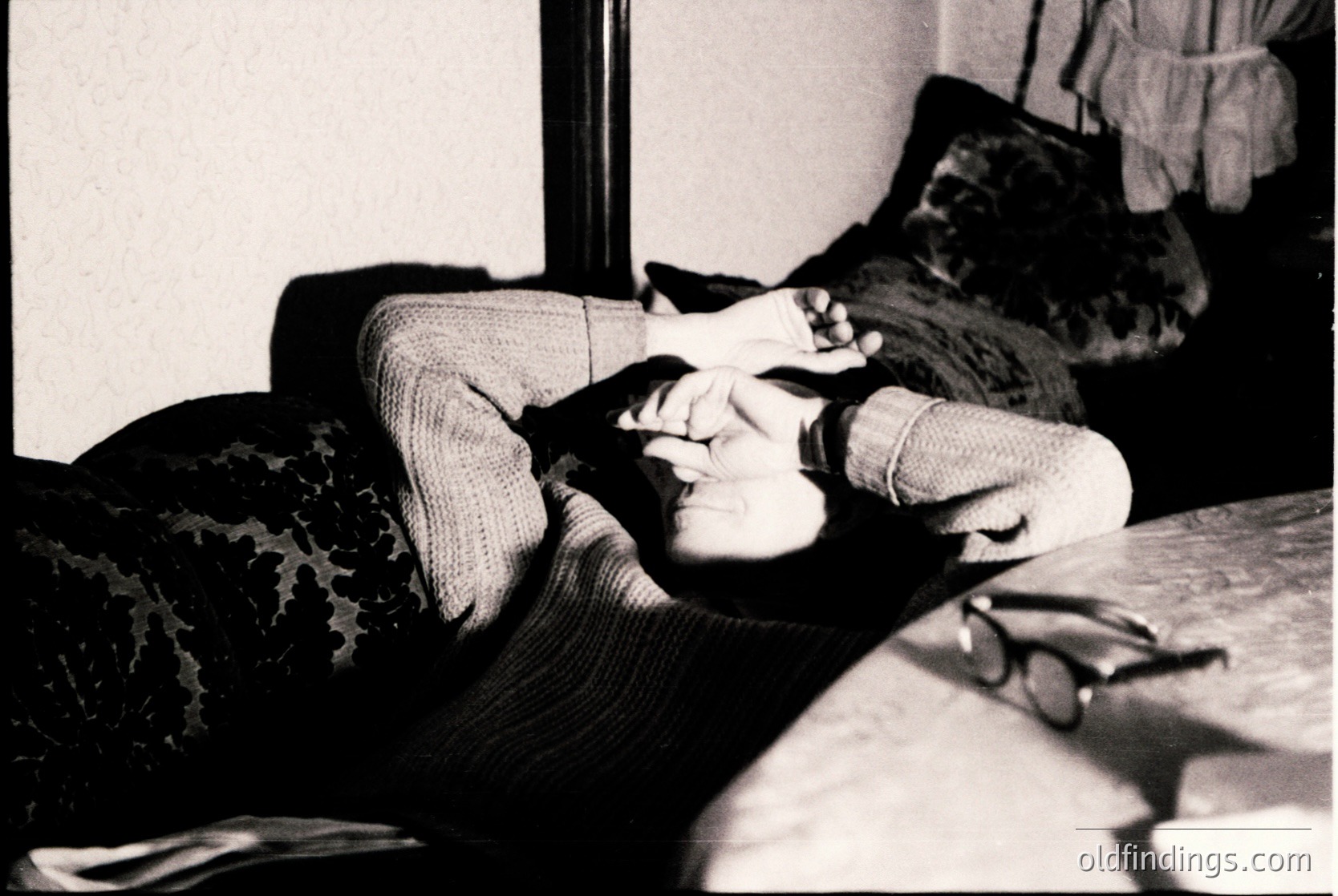 Black-and-white candid shot of a person reclining on a couch, holding a cigarette in one hand while resting their head on the other. Patterned sweater and patterned upholstery suggest mid-20th century domestic interior. Soft focus and natural lighting evoke a relaxed, intimate atmosphere.