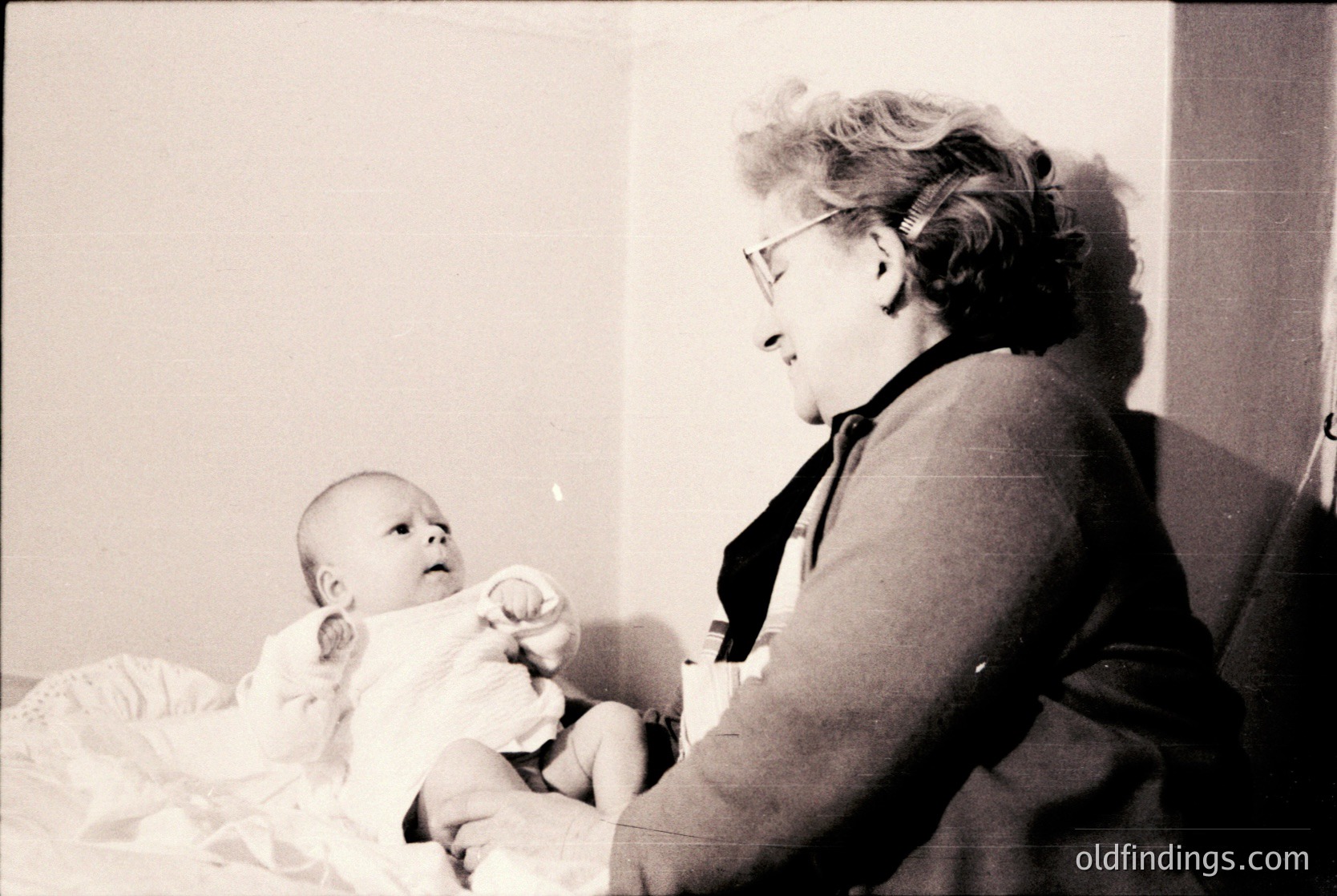 A tender black-and-white moment of a woman in mid-20th-century glasses and a cardigan cradling an infant in a simple room. The baby, swaddled in a light blanket, holds a stuffed toy. Minimalist decor suggests domestic warmth.