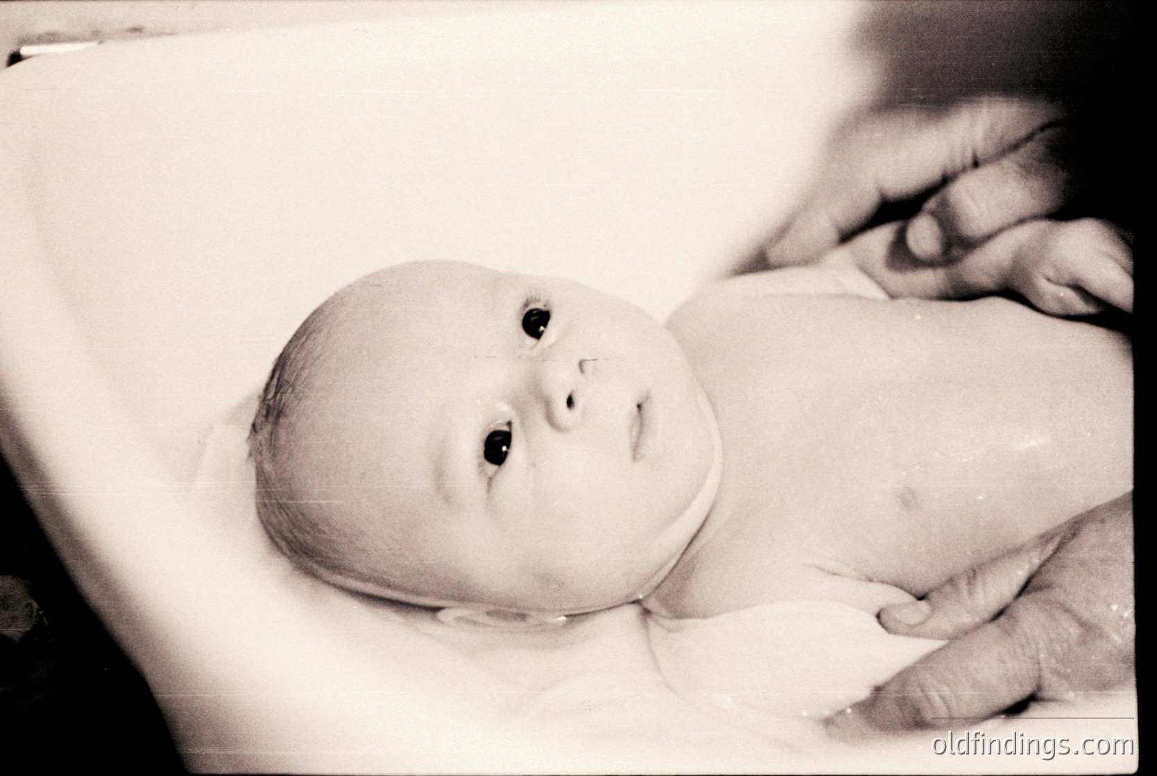 Vintage black-and-white portrait of an infant cradled in an adult’s arms, likely mid-20th century. Soft focus highlights tender moment, with baby’s gaze directed upward. Minimalist composition emphasizes innocence and connection.