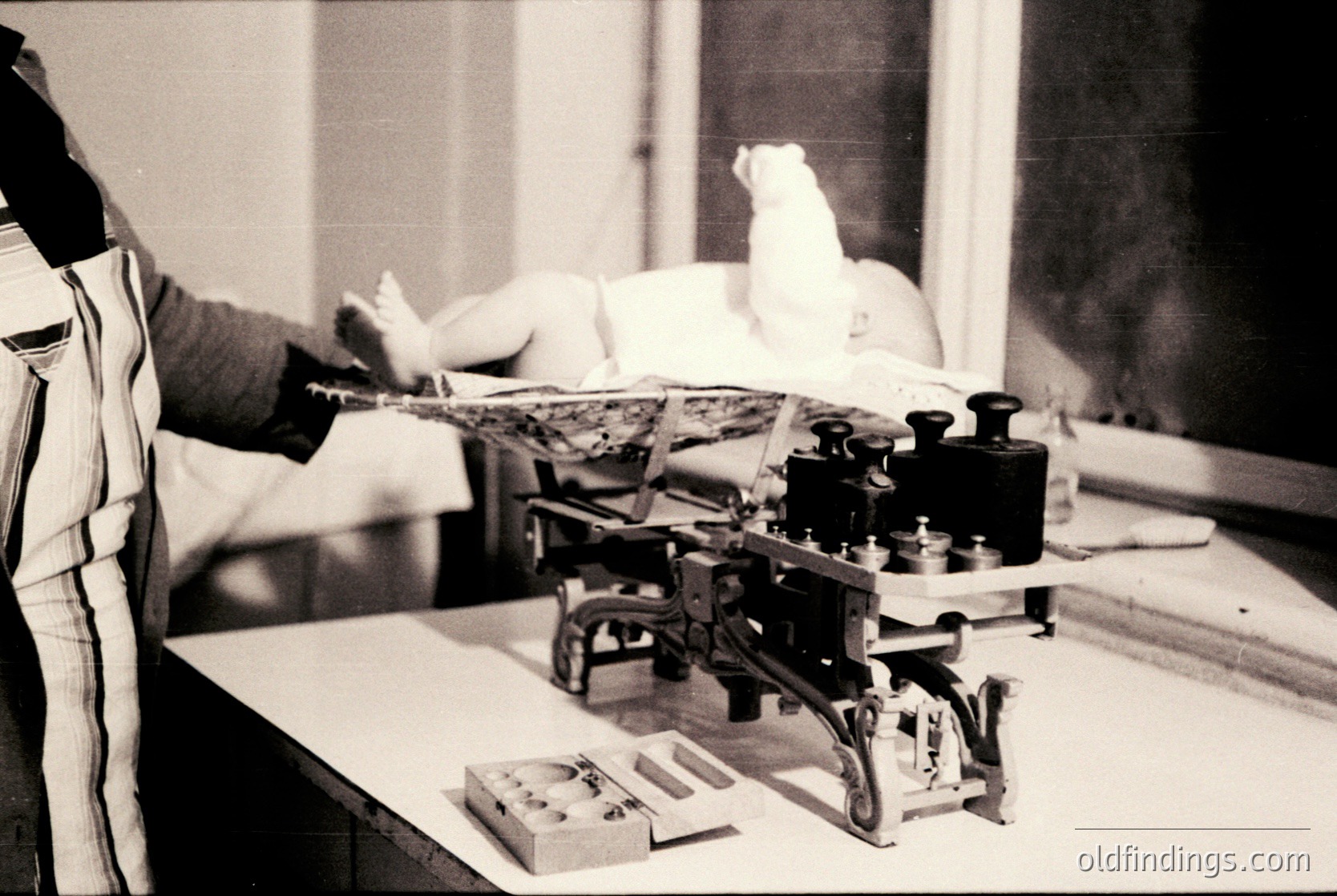 Vintage black-and-white photo of a 19th-century typewriter repair or assembly scene. A person in a striped apron adjusts a manual typewriter with a lever mechanism, while another sits relaxed in a chair beside a window. Industrial-era craftsmanship and manual labor highlighted.