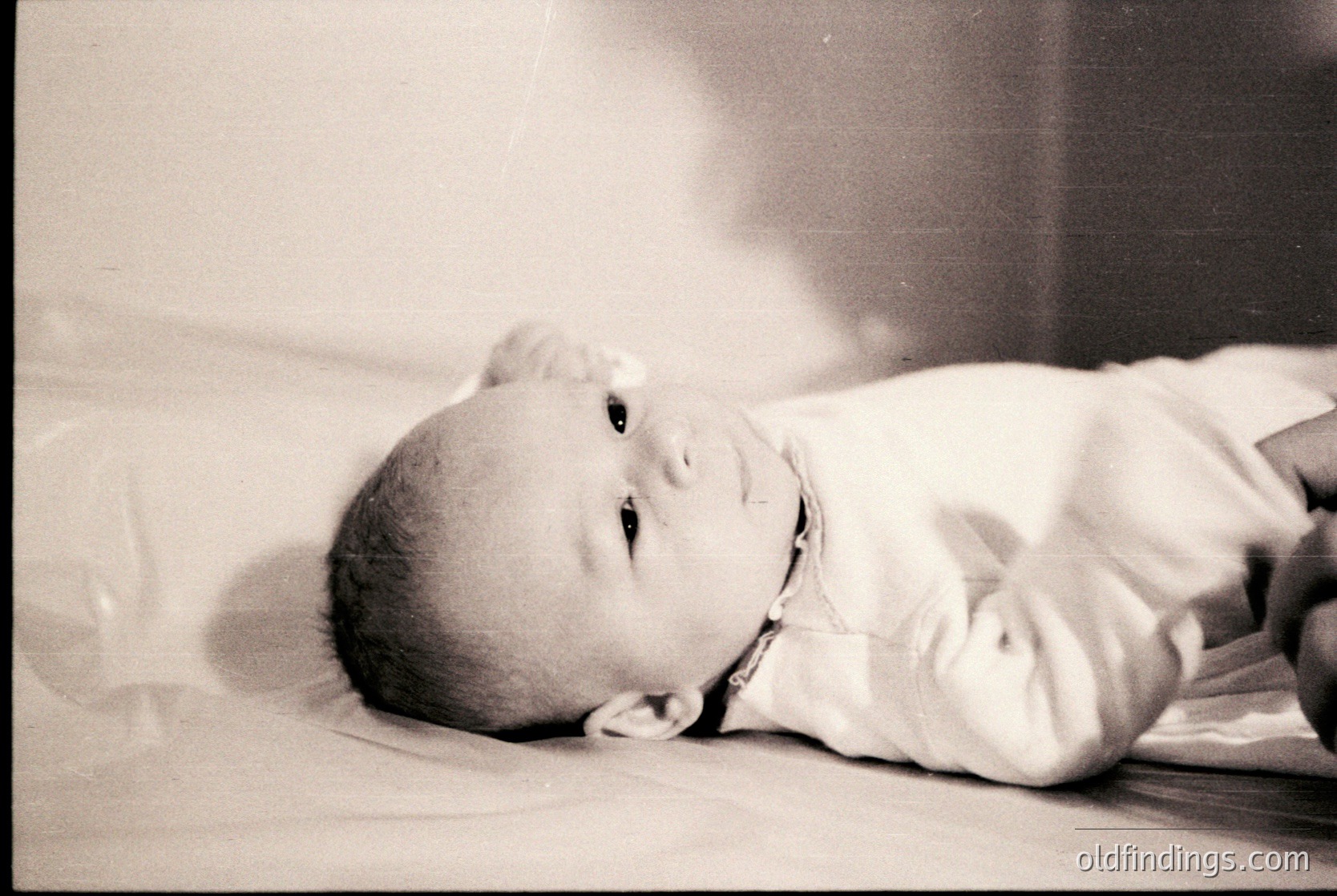 Vintage black-and-white portrait of an infant lying on a bed, dressed in a simple long-sleeve garment. Soft, diffused lighting highlights delicate facial features. Mid-20th century style, likely 1950s–1960s.