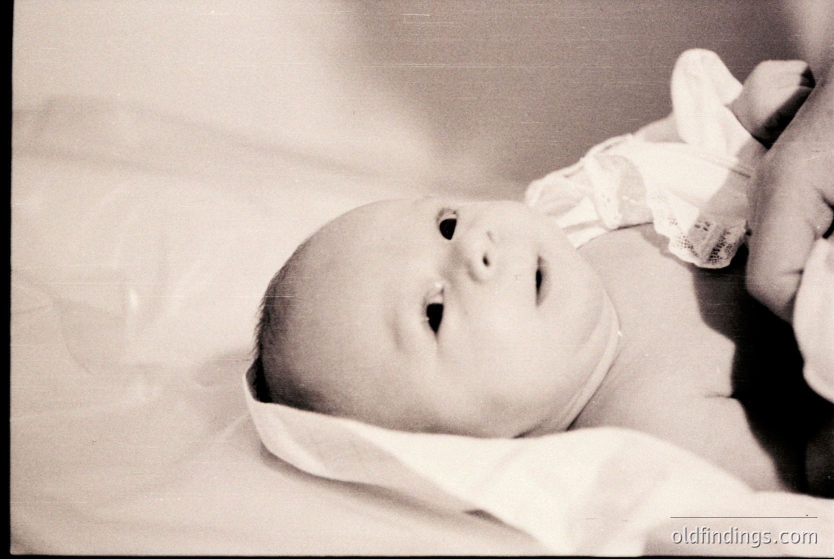 Black-and-white portrait of an infant swaddled in soft cloth, lying on a flat surface. The baby’s expression and open mouth suggest a moment of surprise or curiosity. Classic vintage aesthetic, likely mid-20th century.