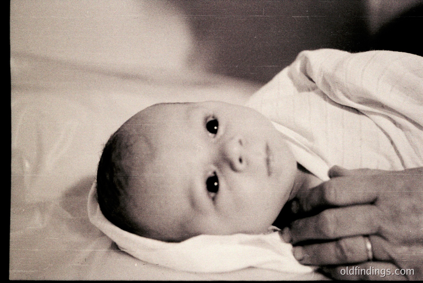 Black-and-white candid of an infant swaddled in a diaper, lying on a flat surface with one hand gently resting on their forehead. Soft focus and vintage grain suggest mid-20th century (1950s–1970s) home photography. Emotional warmth and simplicity evoke mid-century parenting aesthetics.
