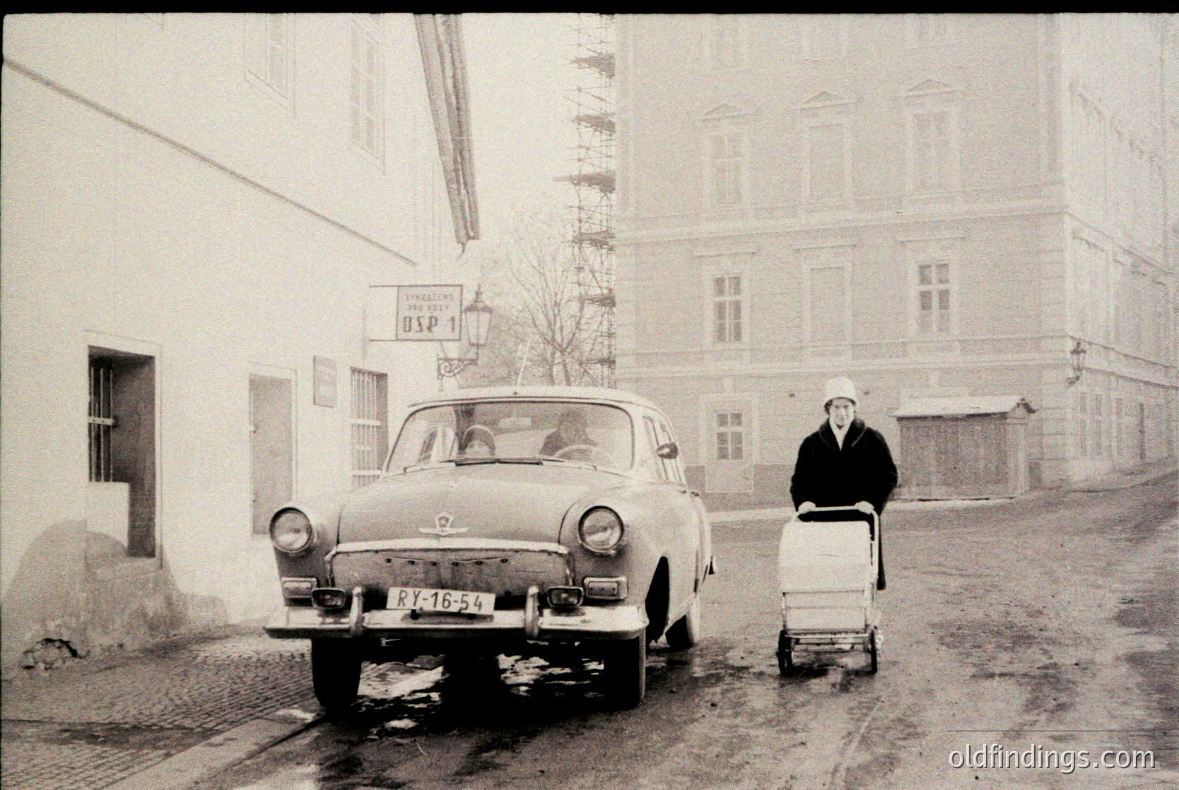Vintage black-and-white street scene featuring a **ZIS-154** (Soviet-era car) parked on a muddy road. A woman in winter attire pushes a white laundry cart, while a man sits in the driver’s seat. Surrounding buildings display Soviet-era signage ("УСП 1") and Soviet-style architecture. Likely **1950s–1960s USSR**.