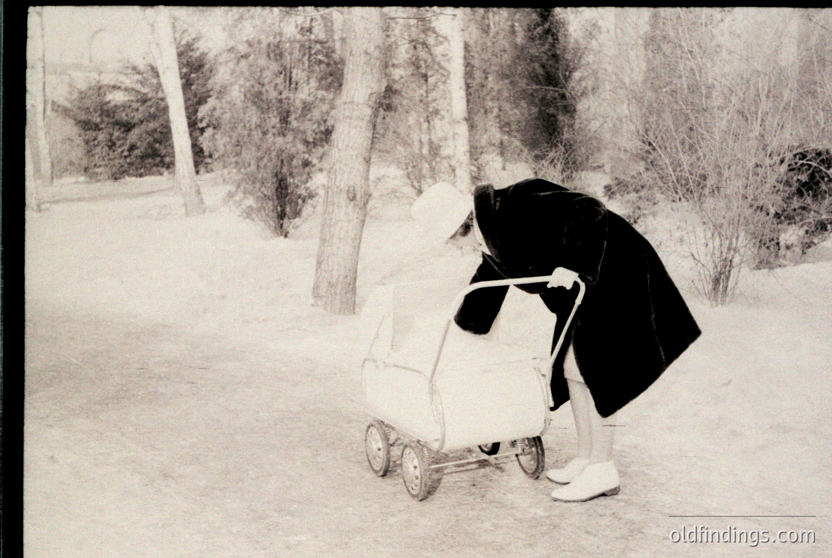 Mid-20th century black-and-white photo of a person pushing a vintage stroller on a snow-covered path. Hooded coat and white snow boots suggest winter attire. Snow-laden trees and bare branches frame the scene, indicating a cold, possibly urban park setting.