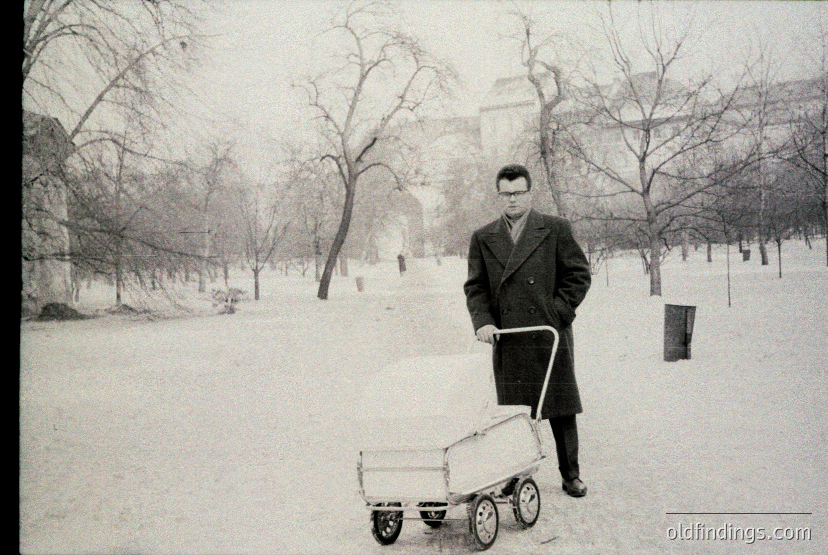 Mid-20th century man in winter coat pushes a wooden wheelbarrow on snow-covered park path, flanked by bare trees. Classic urban park setting with classical architecture in background.