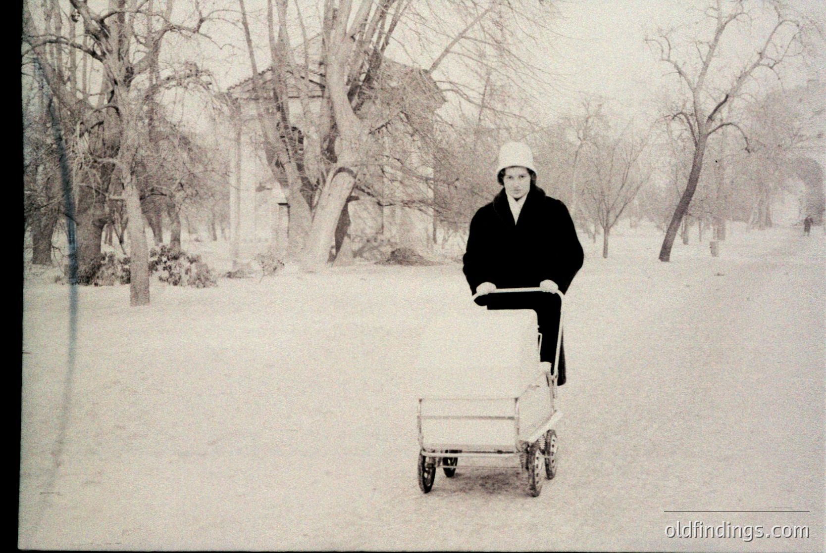 Winter landscape featuring a lone individual pushing a small, open cart through snow-covered park. Leaning coat, white hat, and bare trees suggest mid-20th century urban park setting.
