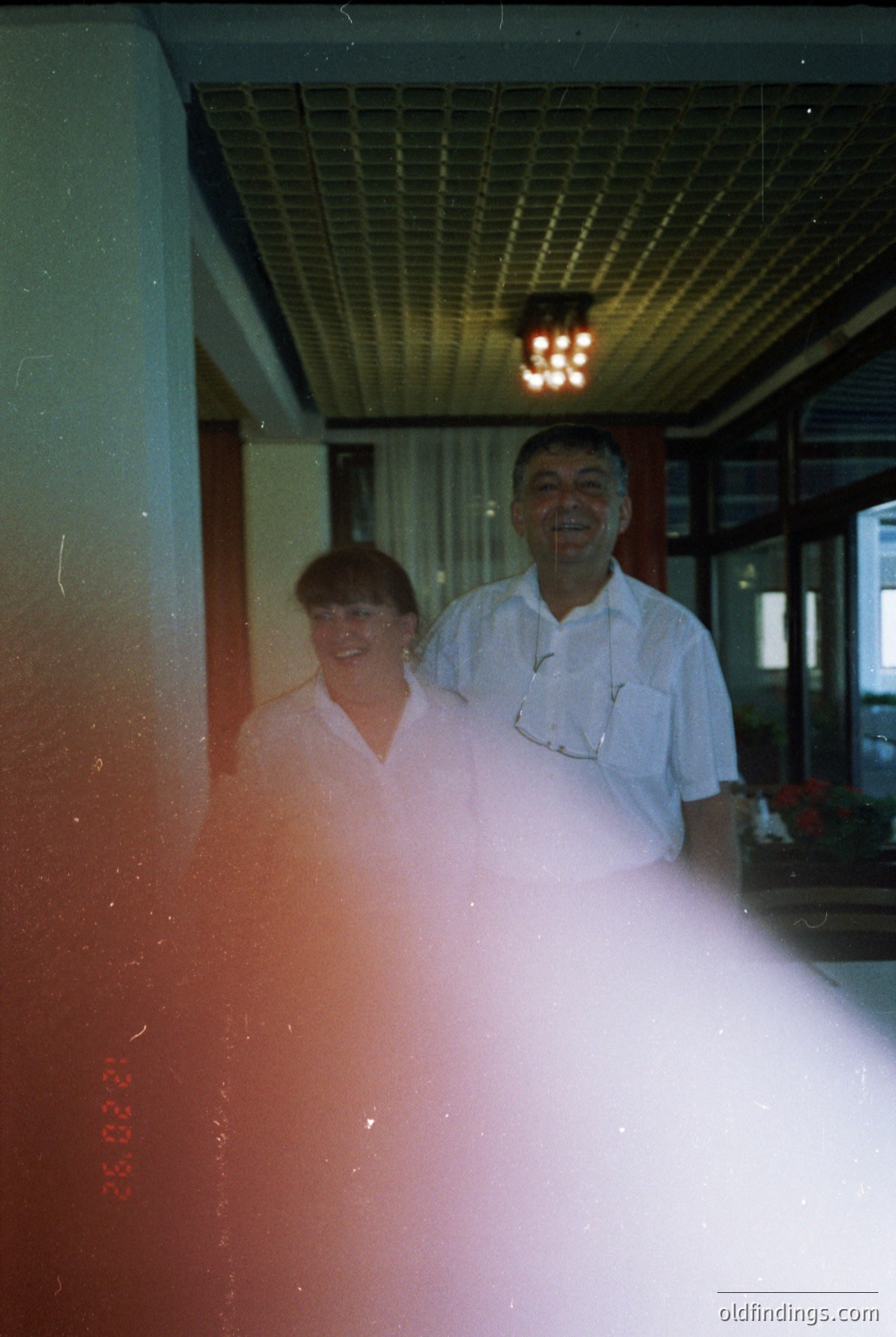 Vintage indoor portrait of two smiling individuals in mid-20th century attire, likely 1950s–1960s. Woman in white blouse, man in white apron with pocket detail. Warm lighting from ceiling fixture, blurred foreground with red tint.