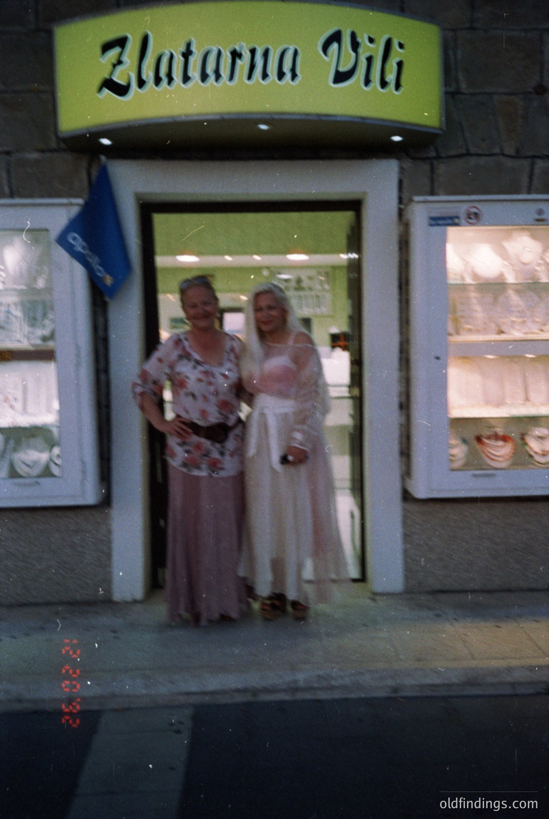 Two women pose outside a vintage jewelry shop, "Zlatarna Vili," in Bulgaria’s 1970s–80s era. The woman on the left wears a floral blouse and patterned skirt; the right, a sheer pink dress with a draped shawl. Display cases behind them showcase gold jewelry. Urban street scene with wet pavement.