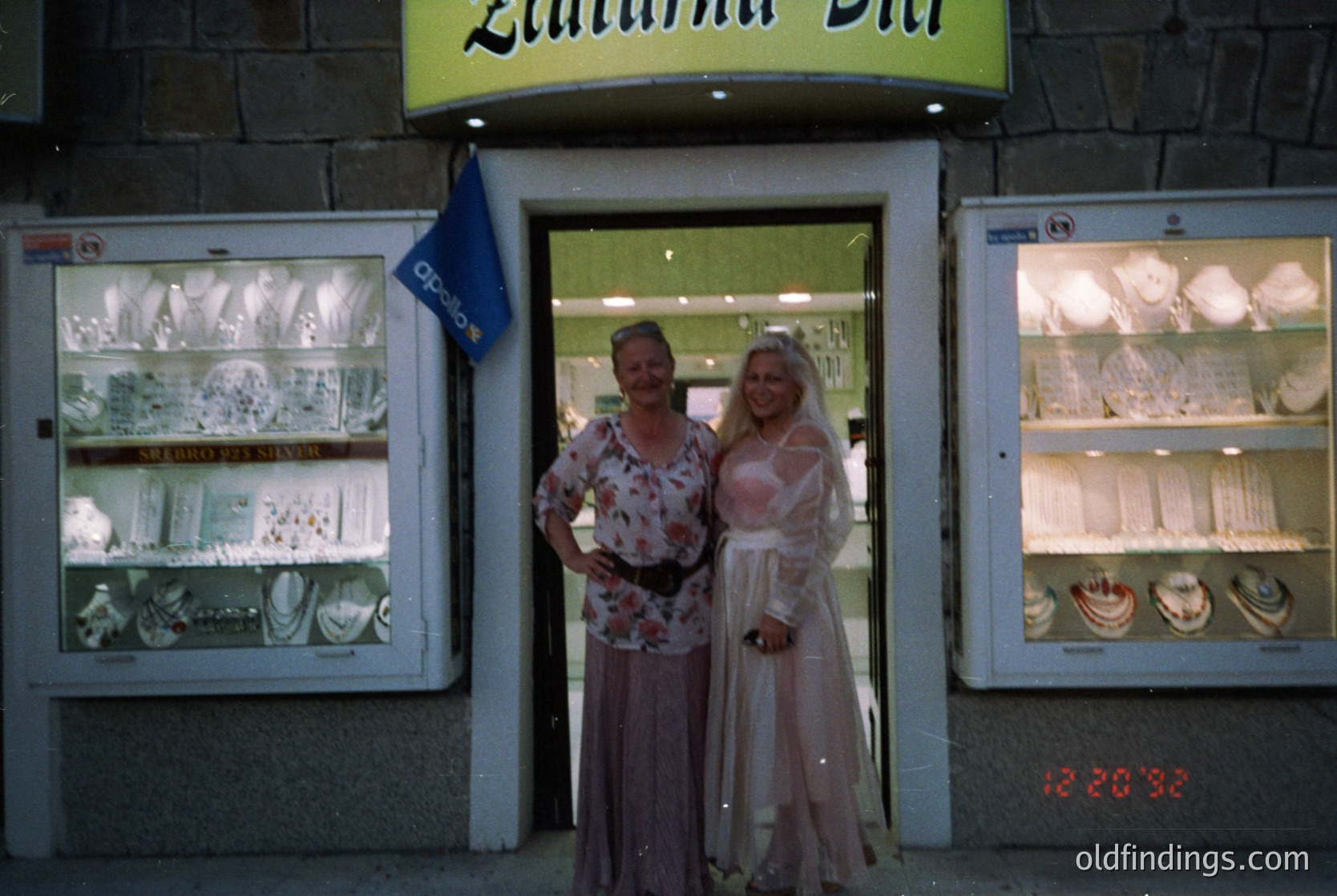 Two women pose outside a vintage silverware shop, "Zlatna Pani," in Bulgaria. The 1970s-era storefront features glass display cases filled with silverware and tableware. The woman on the left wears a floral blouse and skirt; the right, a light pink dress. Neon signage and analog clock (12:20) visible.