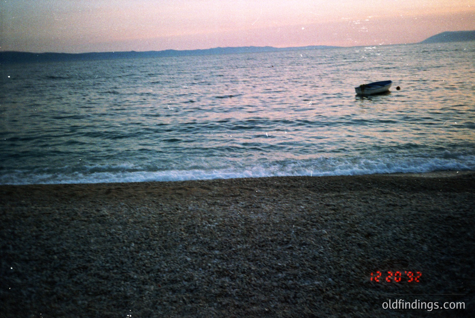 Vintage seaside scene with a small wooden boat anchored near shore at dusk. Waves gently lap against the pebbled beach under a hazy, golden sky. Timestamp "12:30 93" suggests late 1990s. Ideal for nostalgic coastal or maritime collections.