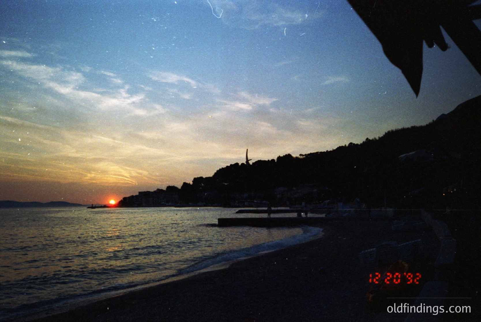 Sunset silhouette over coastal town with illuminated skyline. Low-angle shot captures waterfront buildings, a lone boat, and a clock displaying 12:20:38. Likely Mediterranean or Adriatic region, 1980s-90s film grain evident.