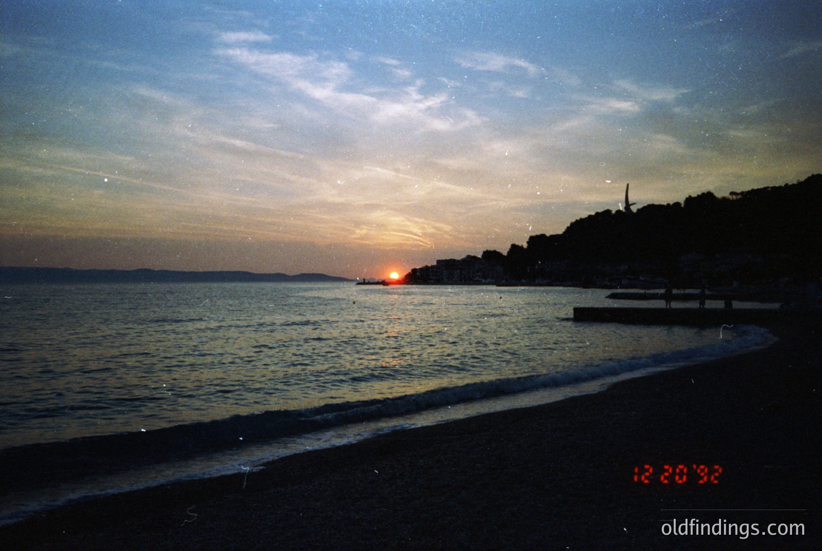 Sunset over a coastal shoreline with silhouetted trees and distant structures. Warm golden light reflects on calm waters, framed by a dark beach. Vintage film grain and timestamp "12.20.95" suggest late 1990s.