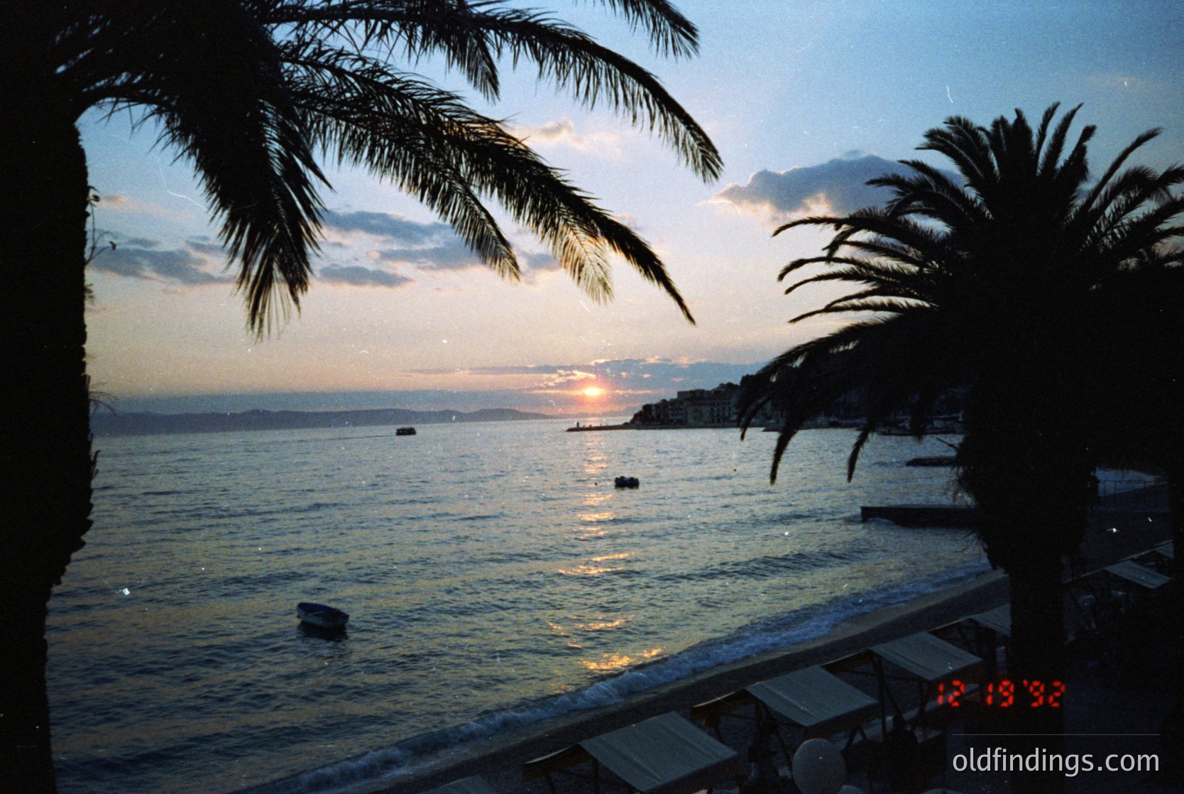 Sunset over a coastal promenade with palm trees framing the view. Warm golden light reflects on calm waters, with silhouetted boats and distant hills. Stamped date: 12/1992.
