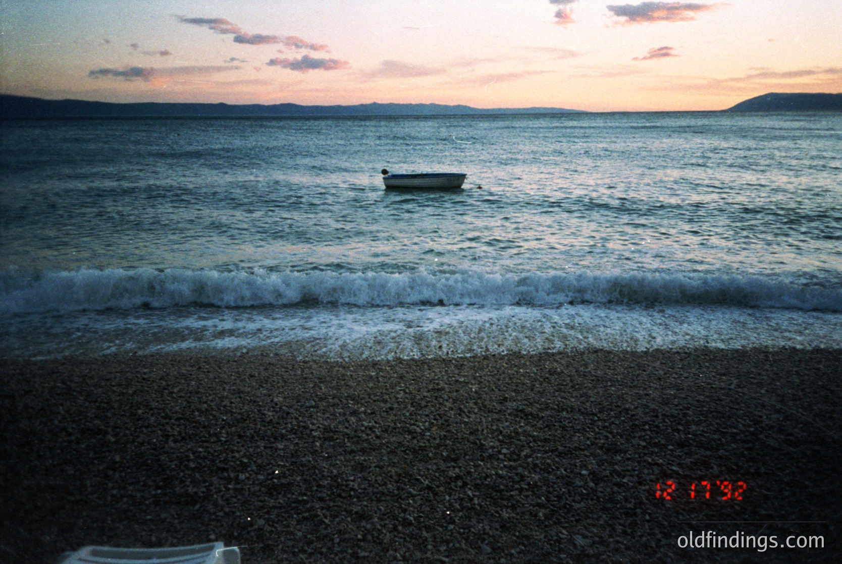 Golden-hour seascape with gentle waves lapping a pebbled shore. A lone wooden boat rests anchored near the horizon, silhouetted against soft clouds and distant land. Vintage film grain and timestamp (12/17/93) suggest late 1990s coastal photography.