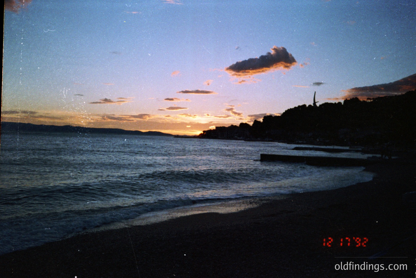 Vintage seaside sunset with golden horizon and scattered clouds over calm waters. Silhouetted trees and distant structures frame the scene. Date stamp "12.17.92" suggests late 1992 coastal shot.