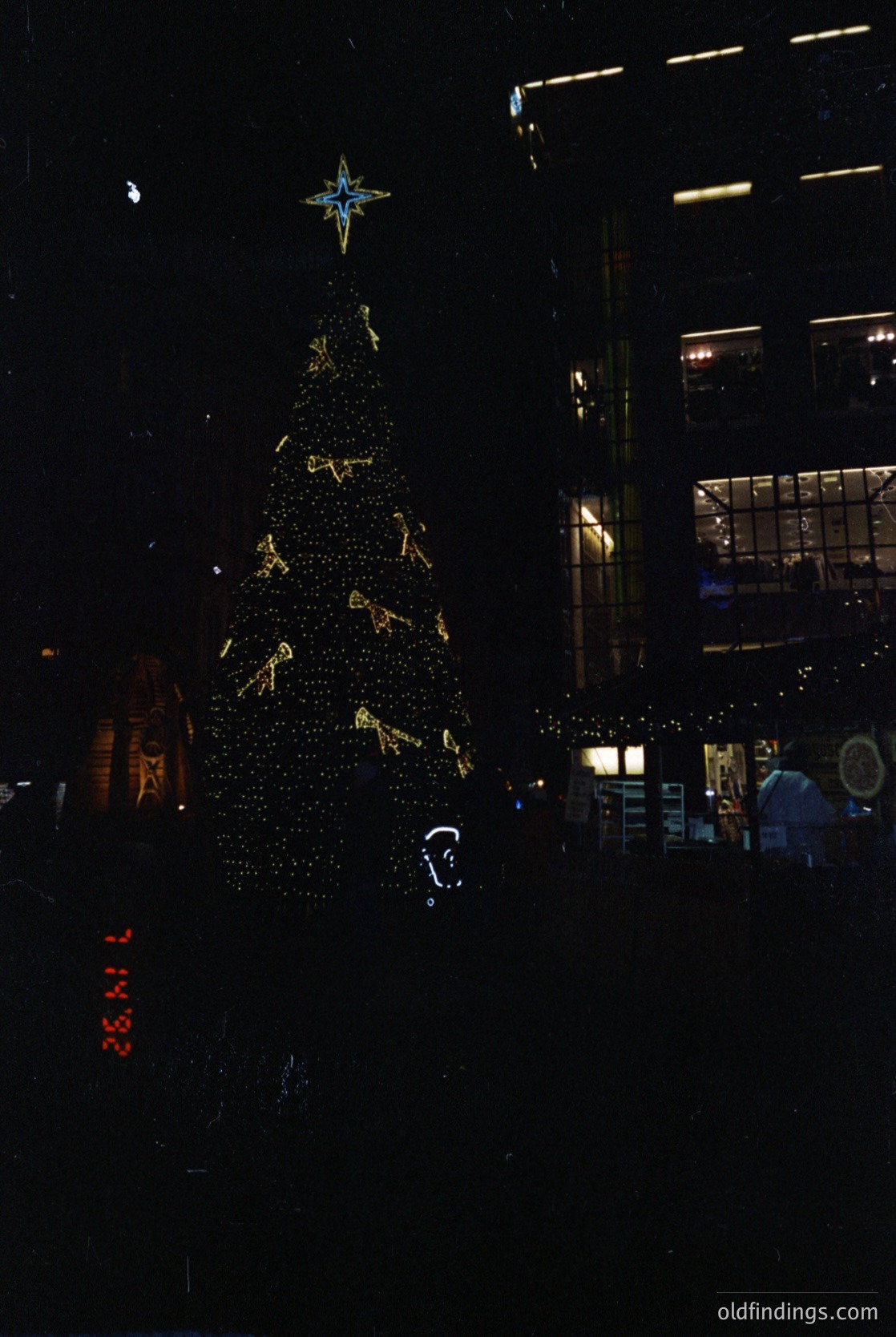 Vibrant nighttime cityscape featuring a towering, illuminated Christmas tree adorned with golden ornaments and a star-topped peak. Surrounding buildings display warm holiday lighting. Urban architecture suggests a European city center. Likely late 20th century holiday display.