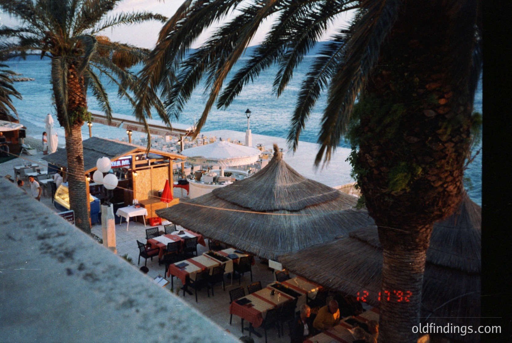 Seaside beachside café with thatched umbrellas and palm-draped tables, overlooking a calm coastal promenade. Architectural details include a mix of concrete structures and natural materials. Likely Mediterranean or tropical coastal region. é