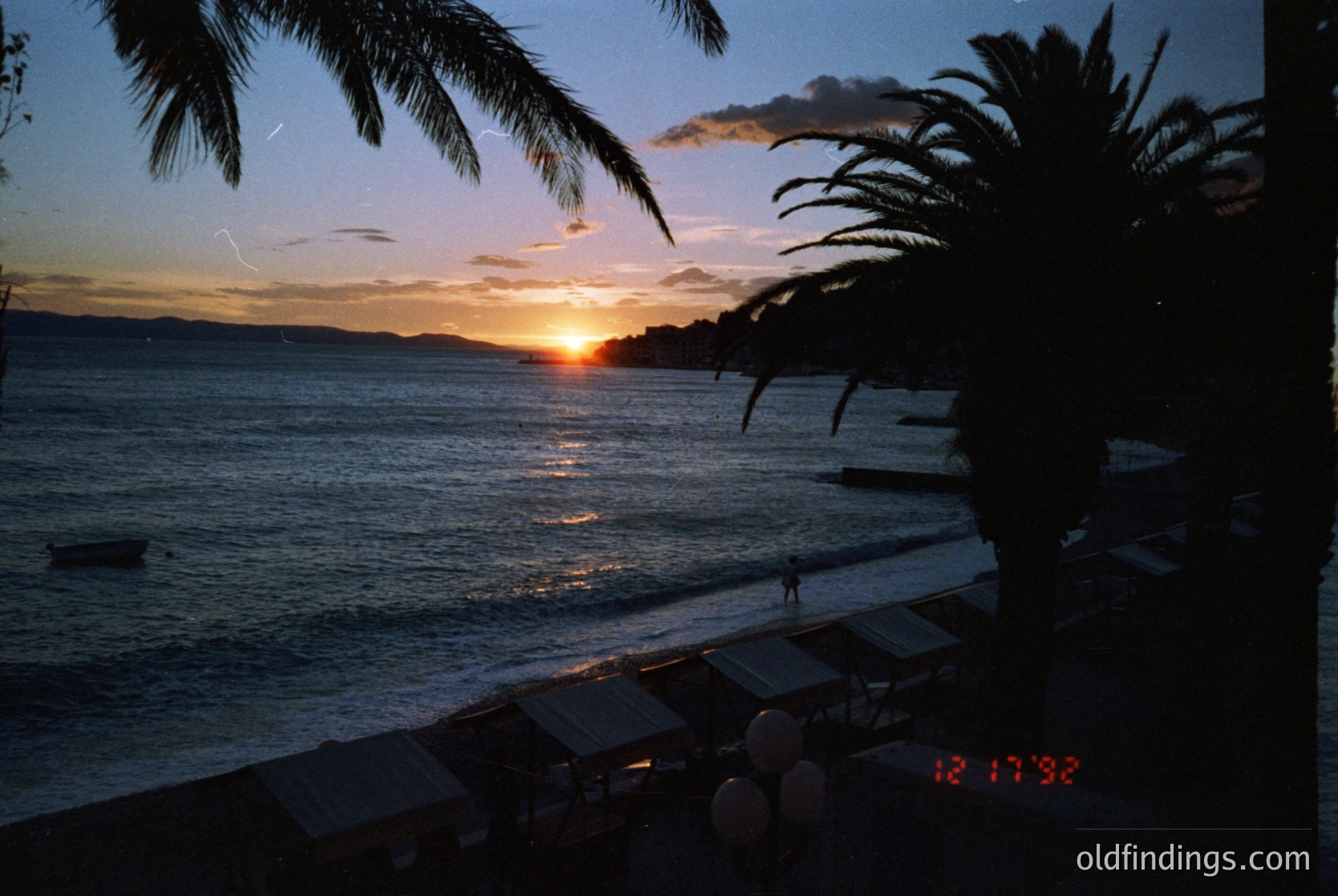Sunset over a calm coastal scene, framed by tropical palm fronds. The digital timestamp (12:17:38) suggests late evening. Empty beach with silhouetted figures near the water’s edge. Warm golden hues reflect on the waves.