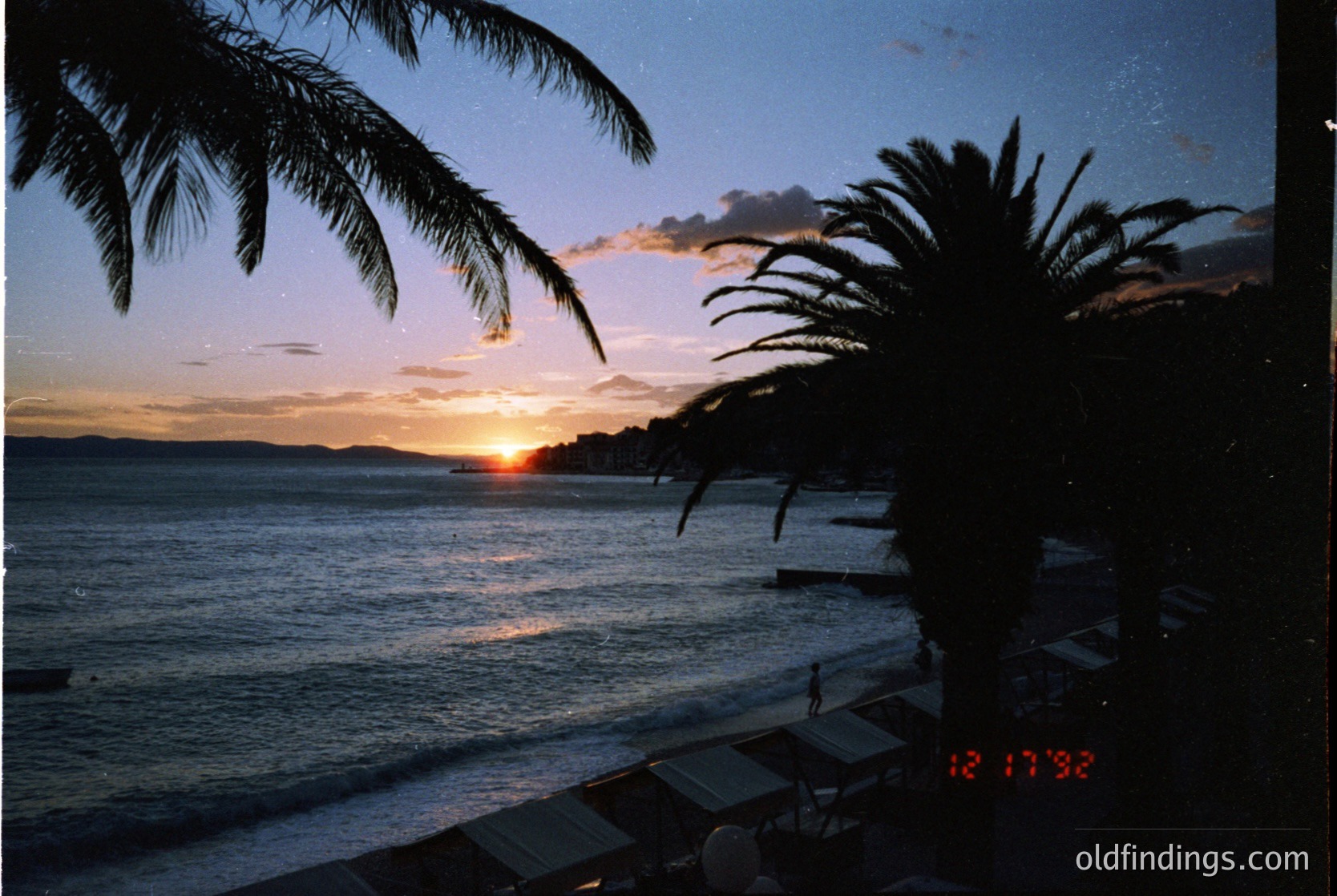 Sunset over a coastal horizon framed by tropical palm fronds, captured 12/17/92. Warm golden light reflects on calm waters, with silhouetted beachgoers and distant boats. Mid-1990s seaside resort ambiance.