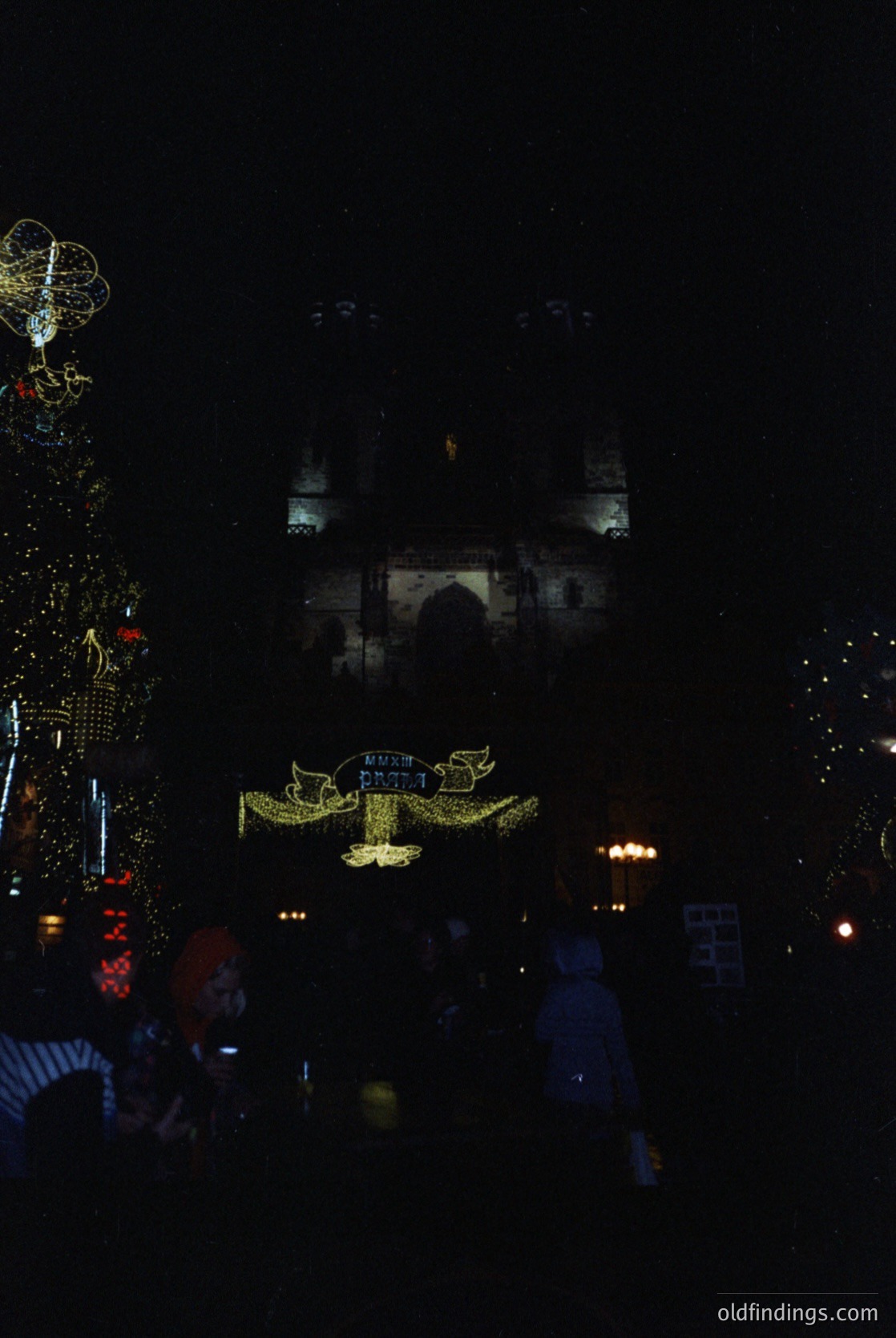 Nighttime festive display featuring illuminated architectural projections on a historic building, likely a mosque. Decorative lights form geometric patterns, including a prominent golden crescent and star motif. Crowd silhouettes suggest a public holiday celebration, possibly Eid al-Fitr or Eid al-Adha. Urban setting with cultural significance.