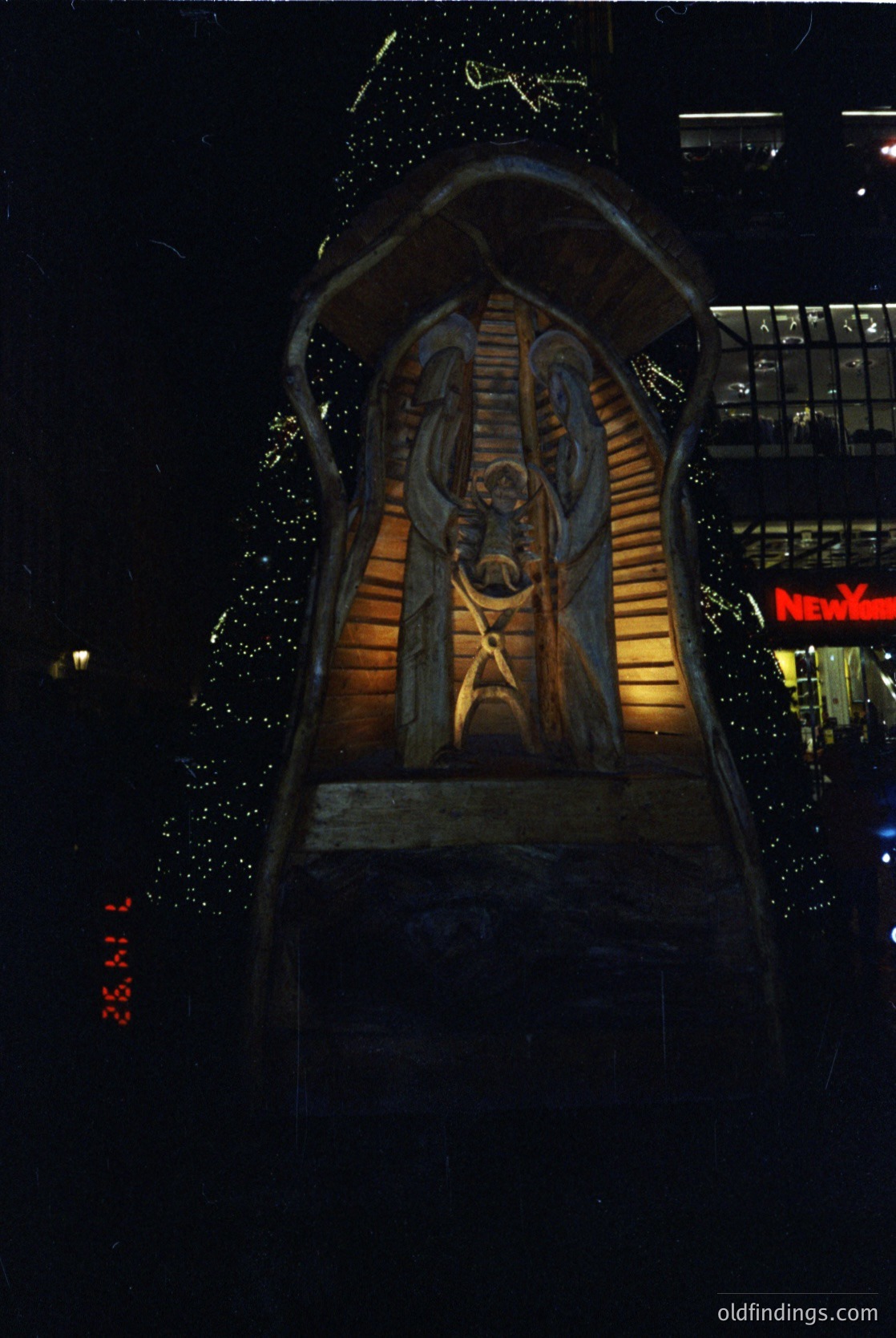 Relief sculpture of a robed figure holding a cross, illuminated at night in an urban setting. The carving features intricate details, including draped fabric folds and a halo. Likely a religious or commemorative monument, possibly from the 20th century. The "26.11" timestamp suggests a vintage or archival photo.