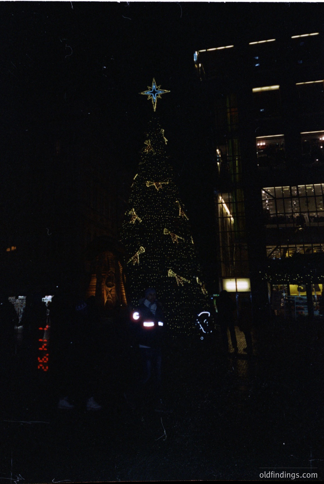A towering, illuminated Christmas tree adorned with golden ornaments and a star-topped crown stands in an urban plaza. Neon signs and modern architecture frame the scene, suggesting a festive holiday display in a city setting, likely late 20th century.