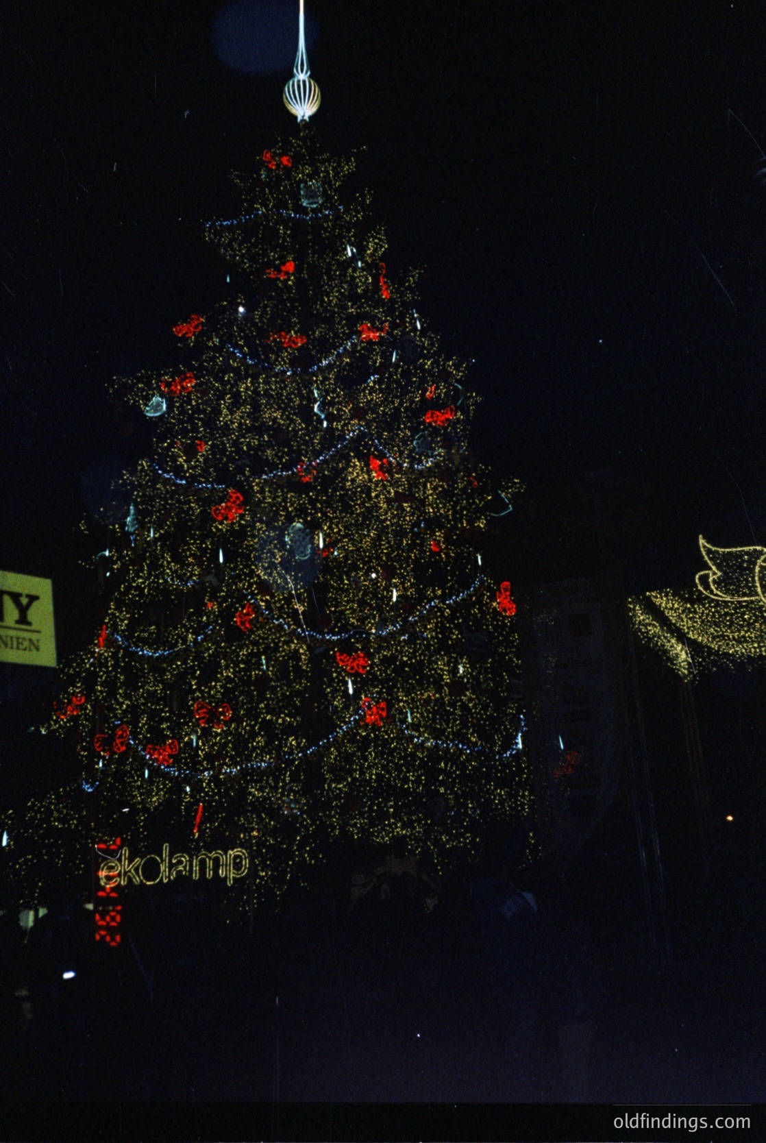 Elaborate gold-toned Christmas tree adorned with red baubles and reflective ornaments, illuminated against a dark backdrop. Branding "ekolamp" visible at base, suggesting a commercial holiday display. Modern, festive design with geometric ornament placement.