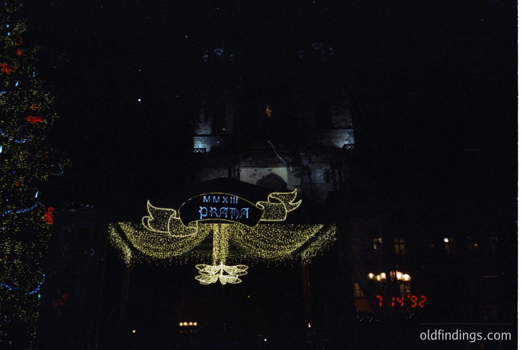 Vintage illuminated banner reading "ПРАЗНИЦА ММХХIII" (Celebration 2003) in Cyrillic, likely for a 2003 anniversary or festival. Decorative golden embellishments and a central floral motif. Urban nighttime setting with blurred city lights and a clock displaying "14:32".