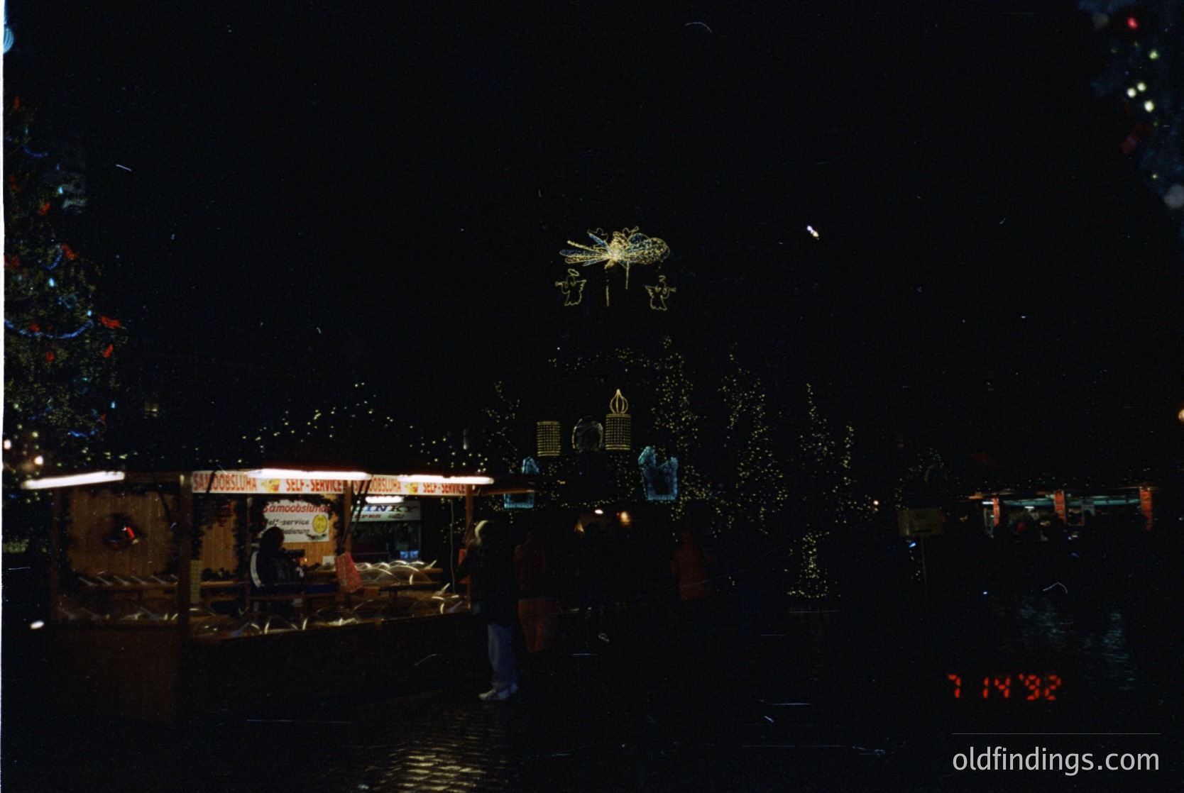 Vintage nighttime street scene with illuminated holiday decorations and a vintage camera timestamp (7:14:38). Decor includes a star-shaped ornament and a golden wreath. A person in dark clothing stands near a small wooden structure with "SOS" signage. Snowflake confetti drifts in the air.