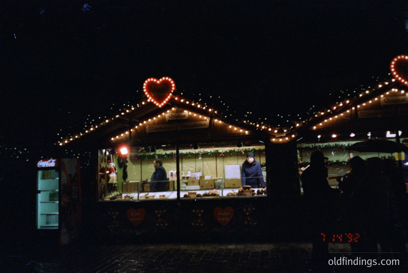 Nighttime market stall adorned with warm string lights and heart-shaped decorations. Indoor seating with blurred figures, likely vendors or patrons. Coca-Cola vending machine visible on left. Digital timestamp reads 7:14 PM.