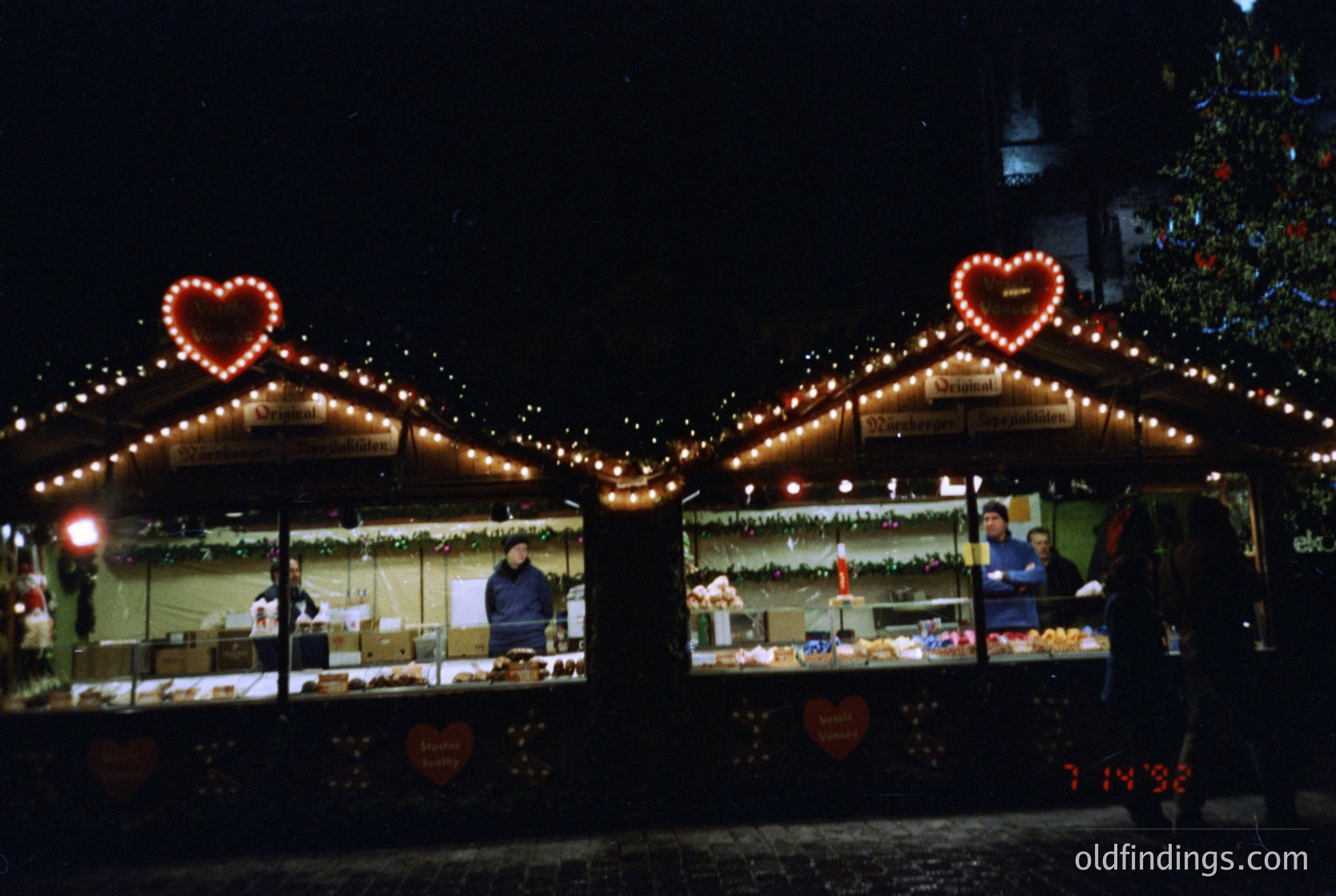 Christmas market stalls adorned with heart-shaped string lights, illuminated at night. Vendors behind counters display baked goods and holiday treats. Decorated pine tree visible in background. Likely European winter festival setting.