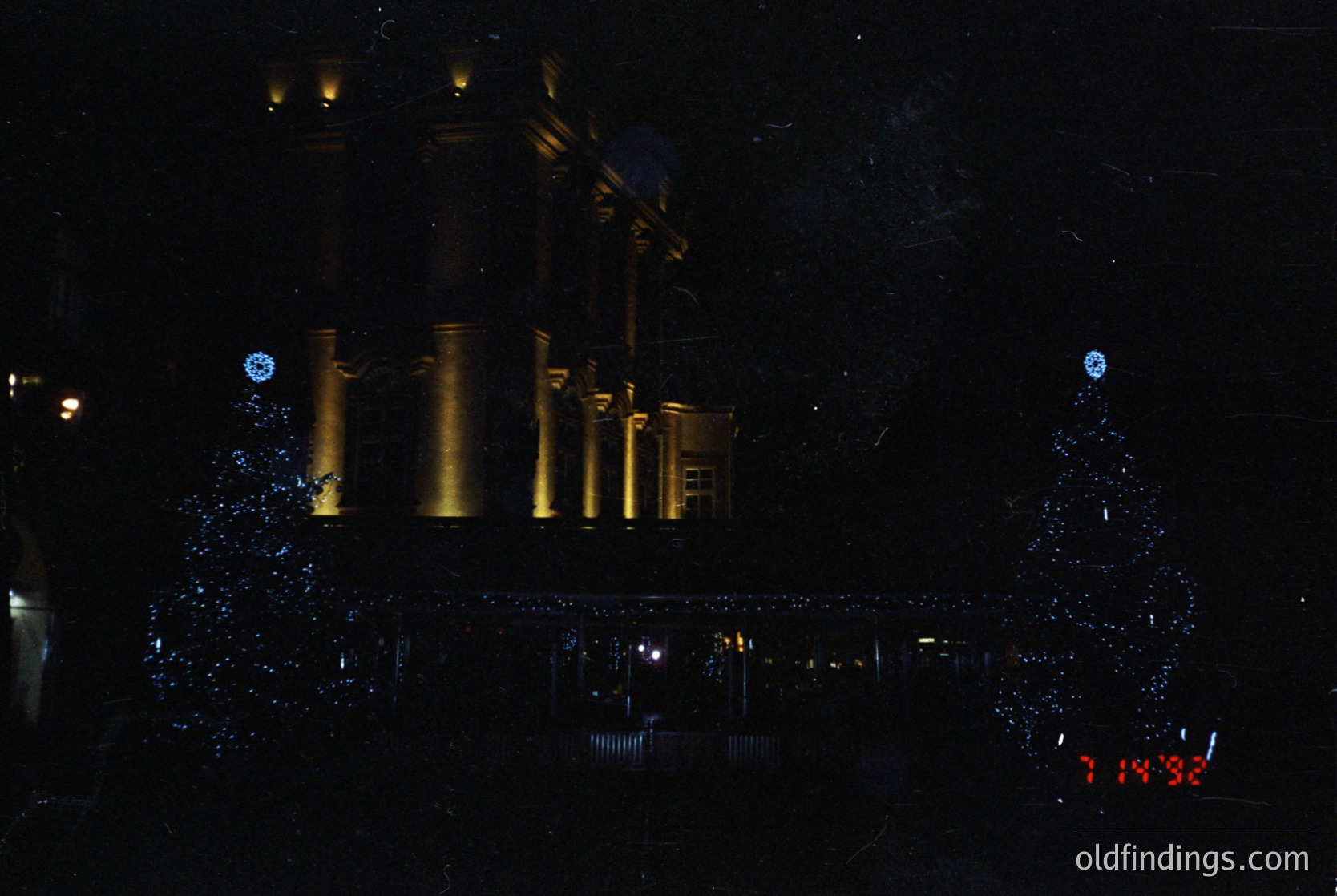 Neon-lit industrial facade at night, featuring four vertical cylindrical tanks with illuminated yellow-orange outlines. Reflections of lights create a mirrored effect on a wet surface below. Time stamp "1:18:32" visible in lower-right corner.